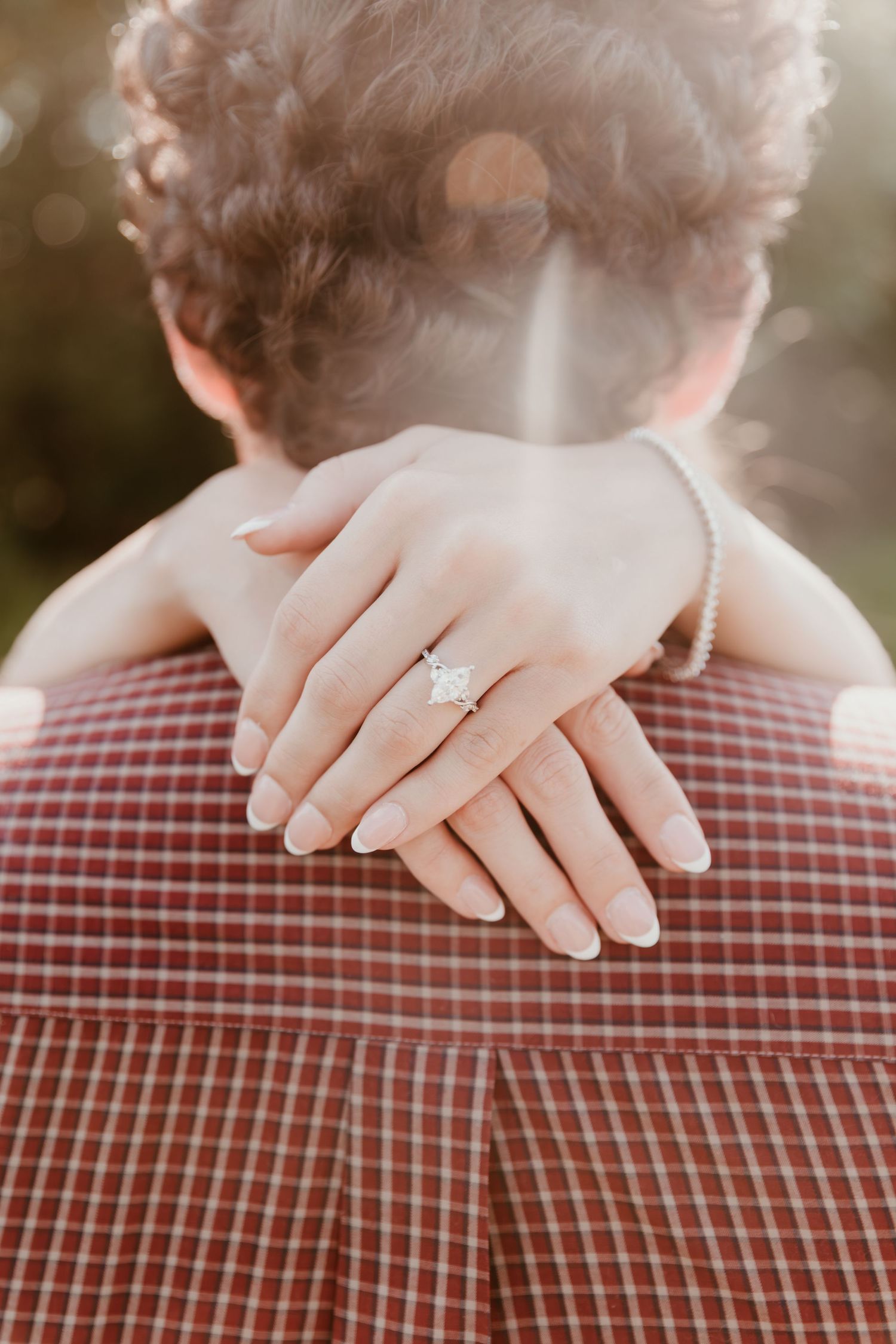 A close-up engagement photo showing hands with a diamond ring resting on a red and white plaid shirt in warm sunlight.