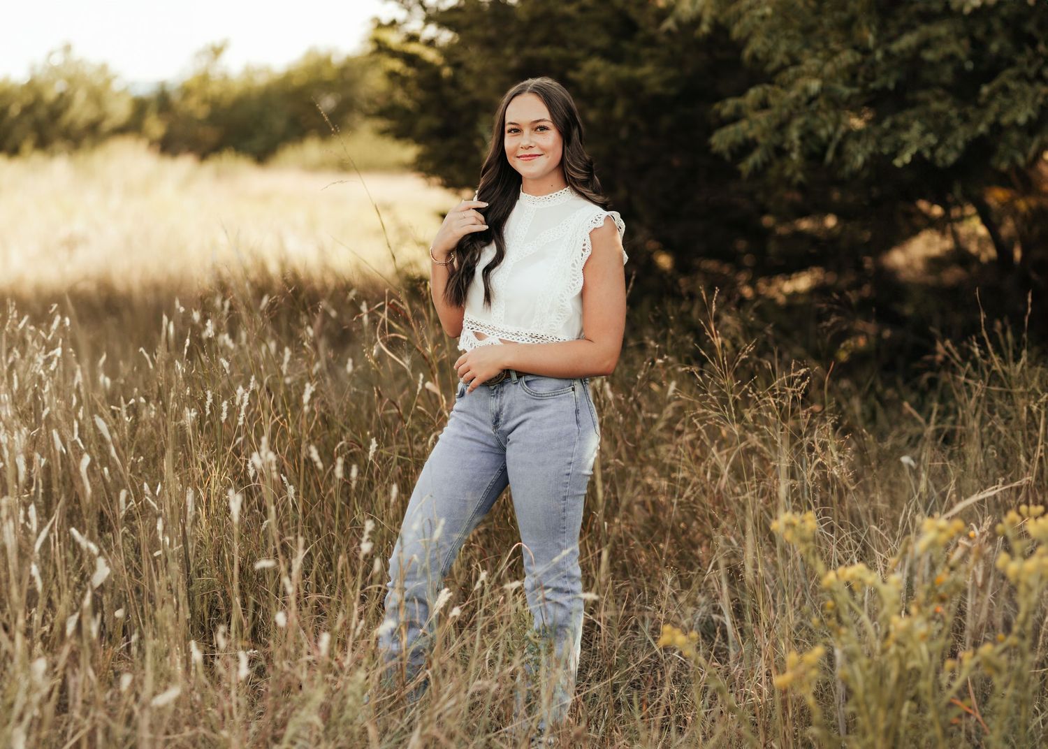 Beautiful outdoor portrait in a golden field with tall grass and evergreen trees at sunset.