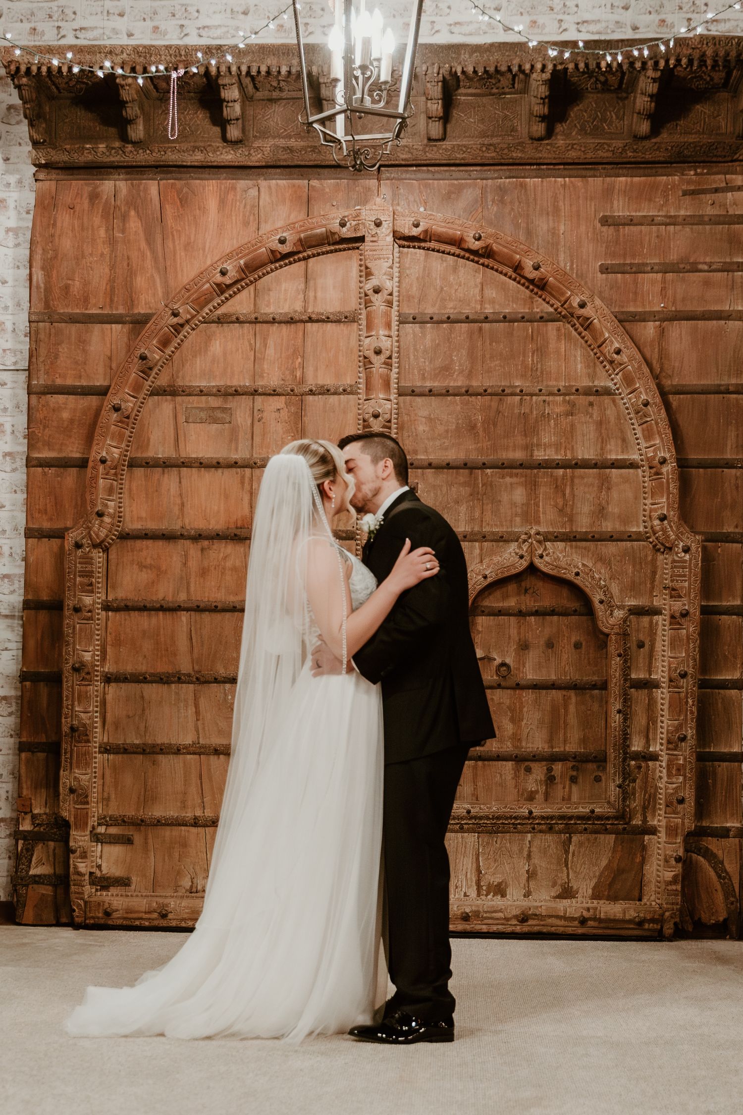 A couple shares a tender moment in front of a rustic wooden arched door with ornate metal details.