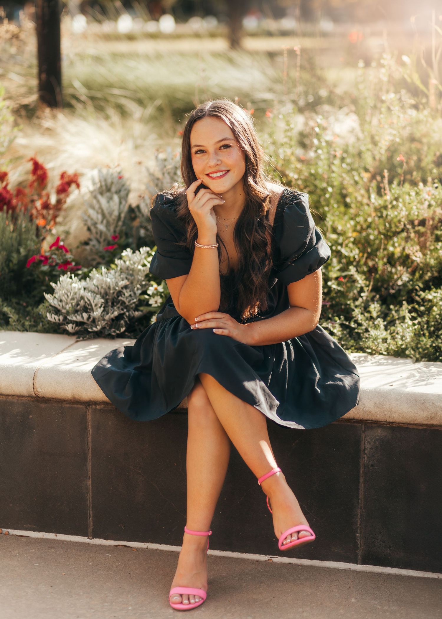 A woman in a navy dress sits on a stone wall surrounded by flowers in warm sunlight.