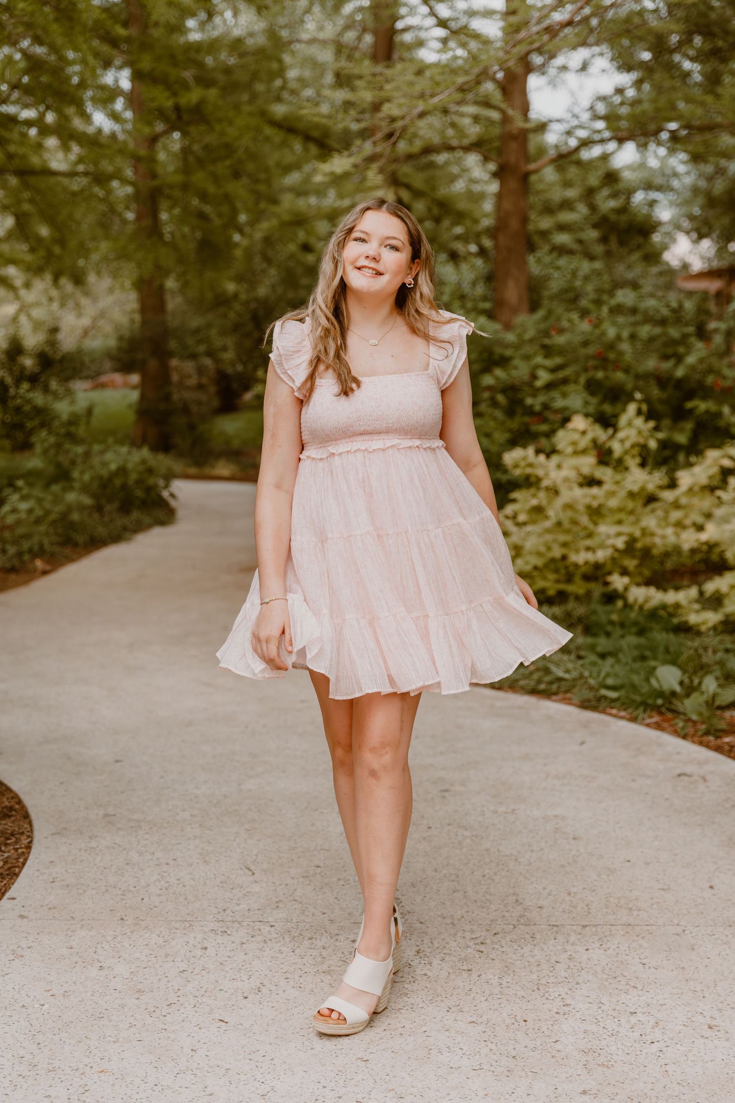 A joyful figure in a light pink summer dress walks along a concrete path surrounded by lush green trees.