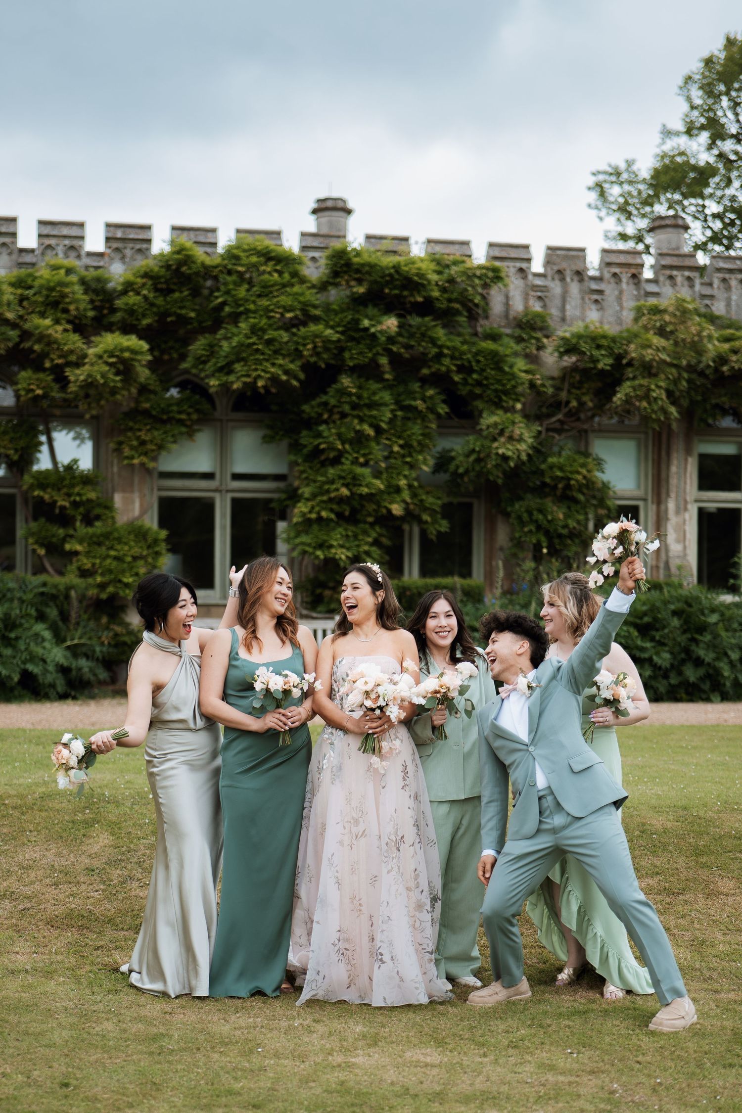 Wedding party celebrates joyfully on a lawn in front of an ivy-covered stone building with bridesmaids in sage and silver gowns.