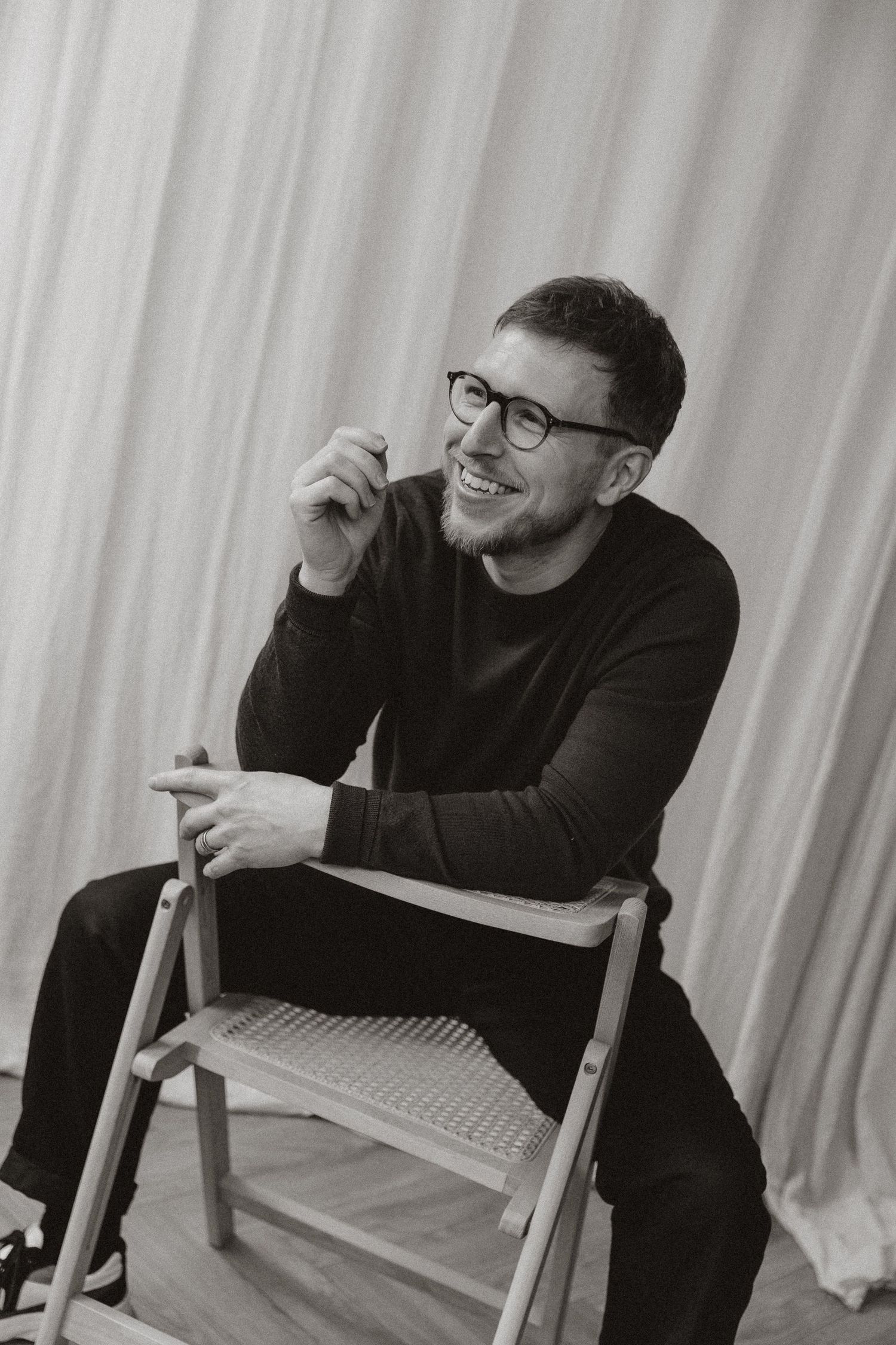 Black and white photo of someone in glasses and dark sweater sitting on chair and laughing.