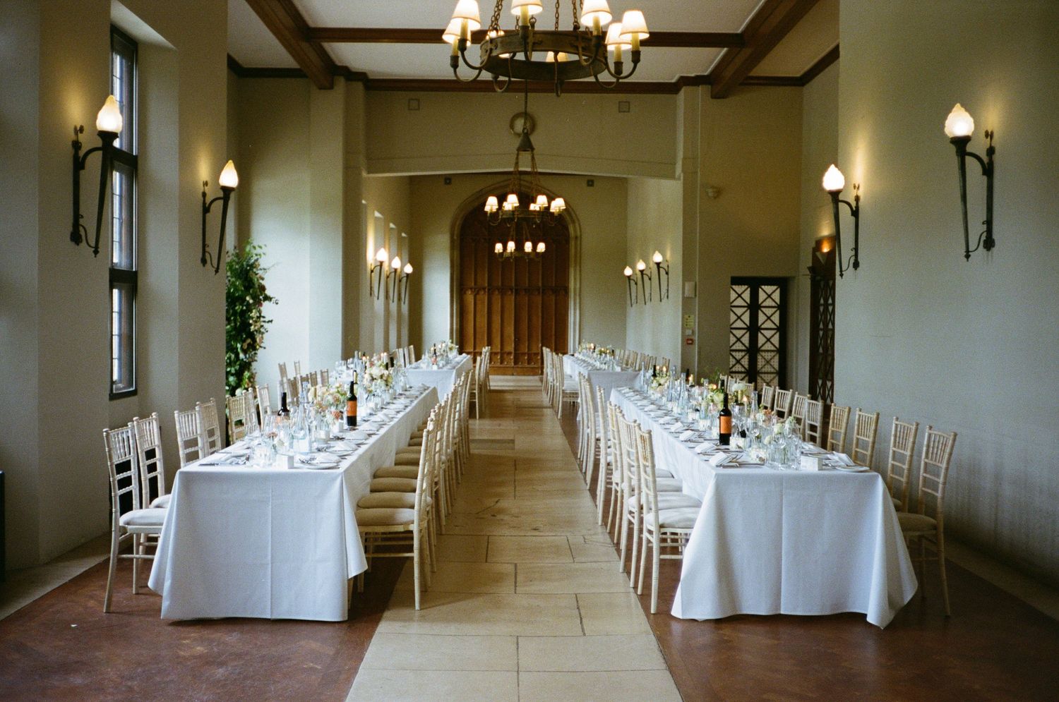 White tables and chairs line an elegant ballroom with tall ceilings, wall sconces, and grand wooden doors at the end.