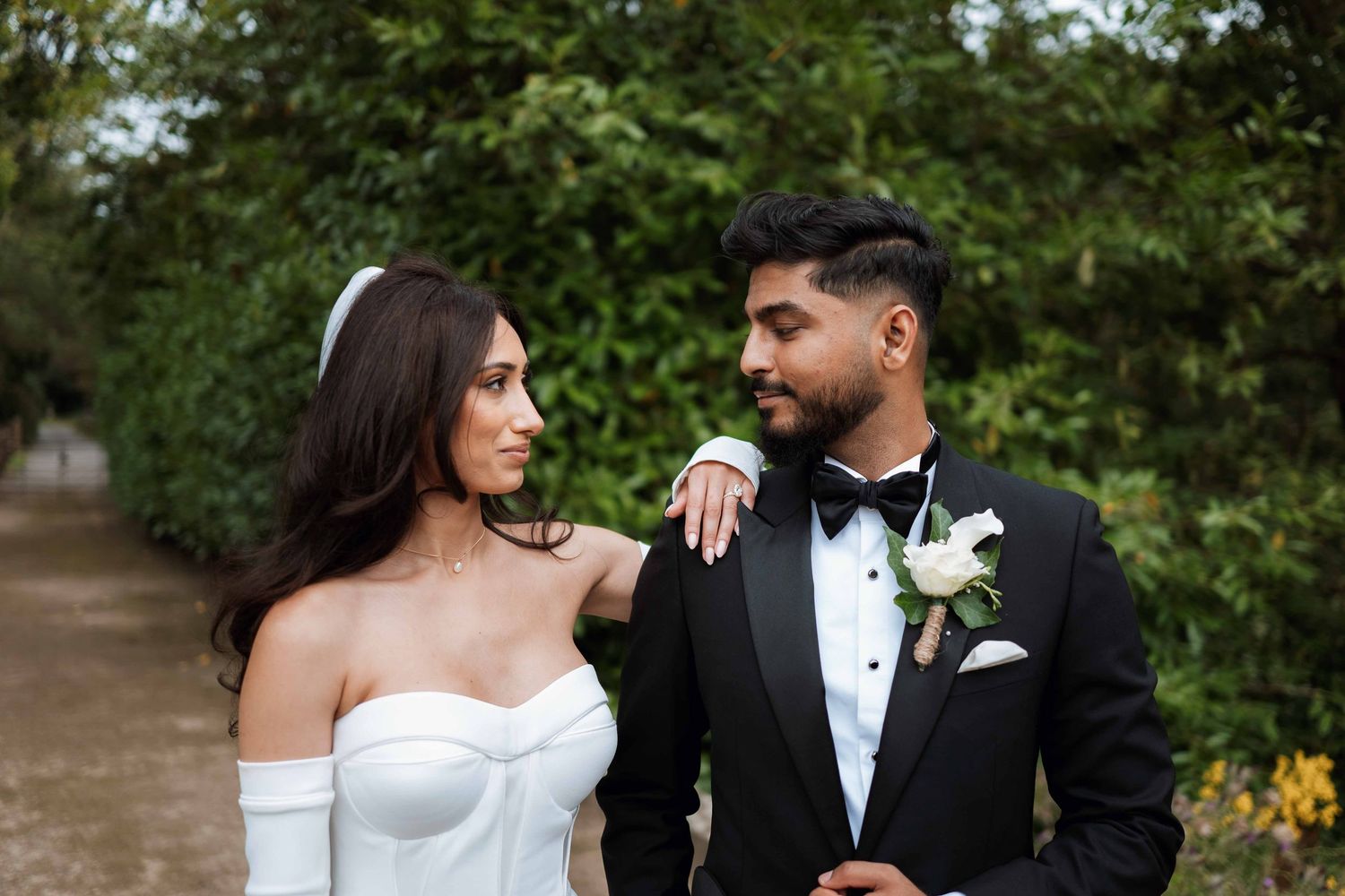 A romantic wedding couple smiles at each other on a garden path, with the bride in white and groom in black tuxedo.