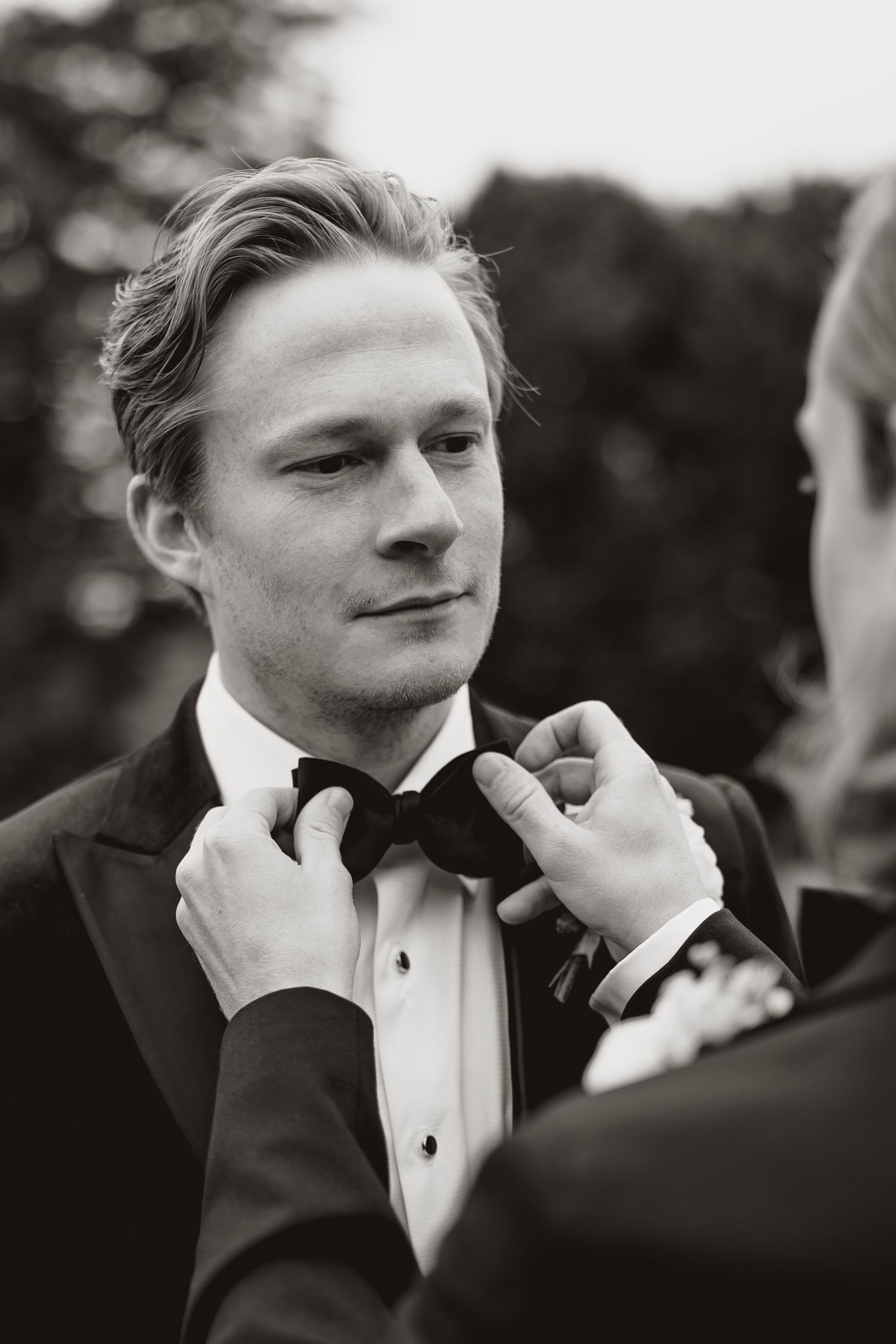 Black and white photo of groom in tuxedo getting bow tie adjusted during wedding preparations.