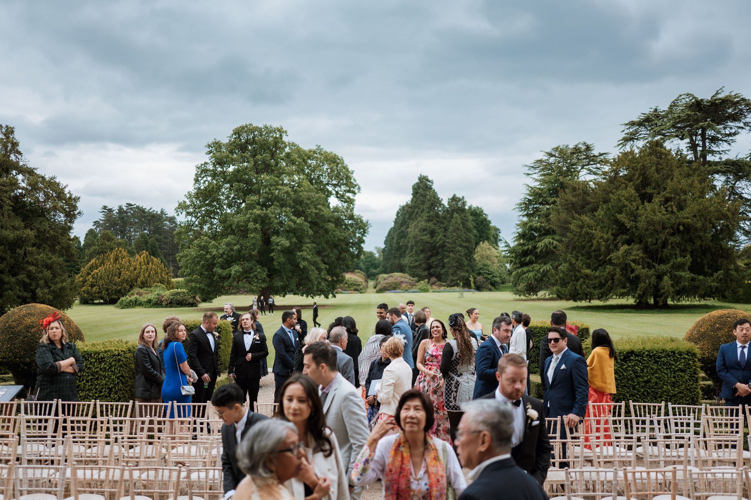 Wedding guests gather on a lush green lawn at an outdoor venue with rows of empty chairs and cloudy skies above.