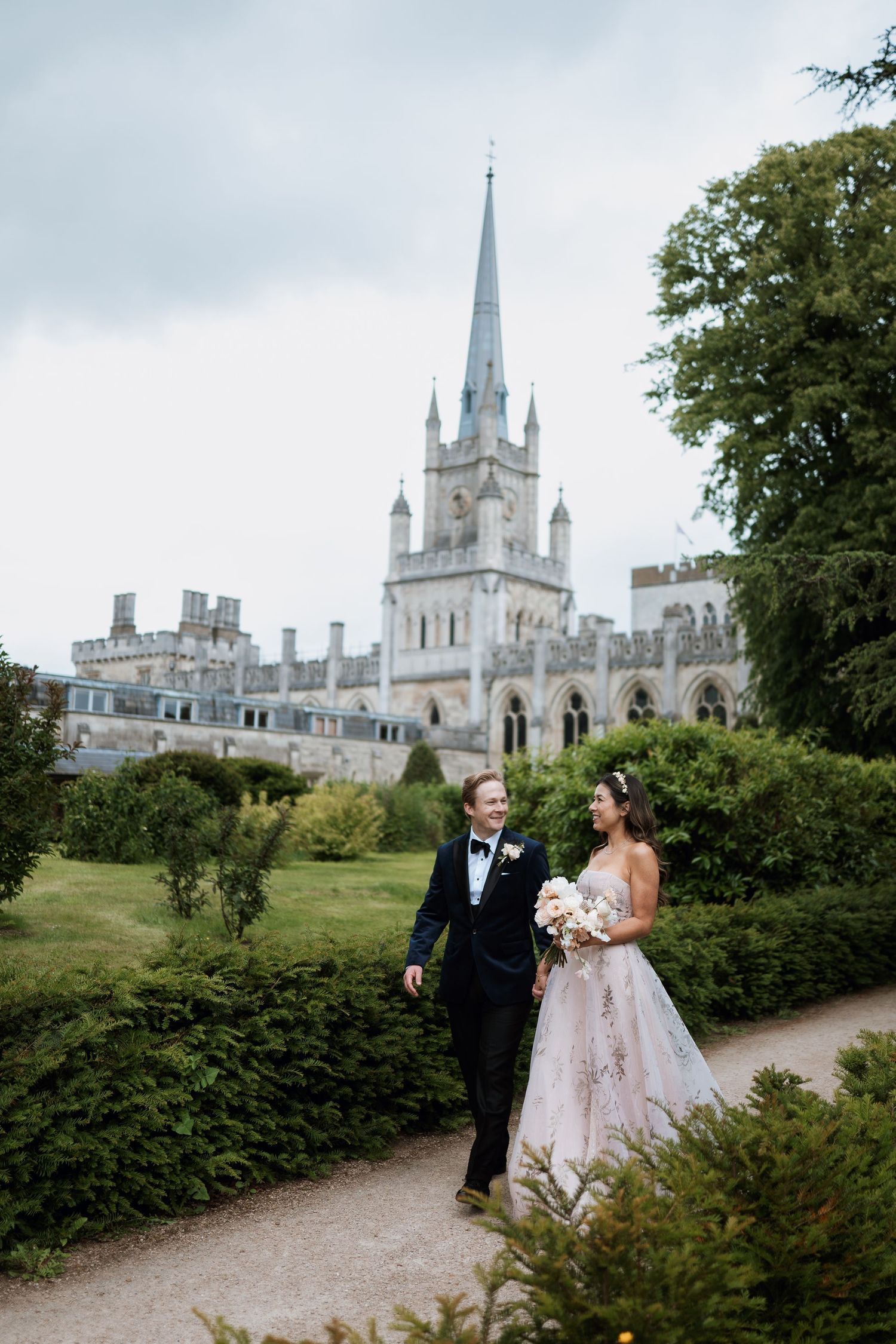 A couple walks along a garden path with a grand Gothic castle featuring a tall spire in the background.