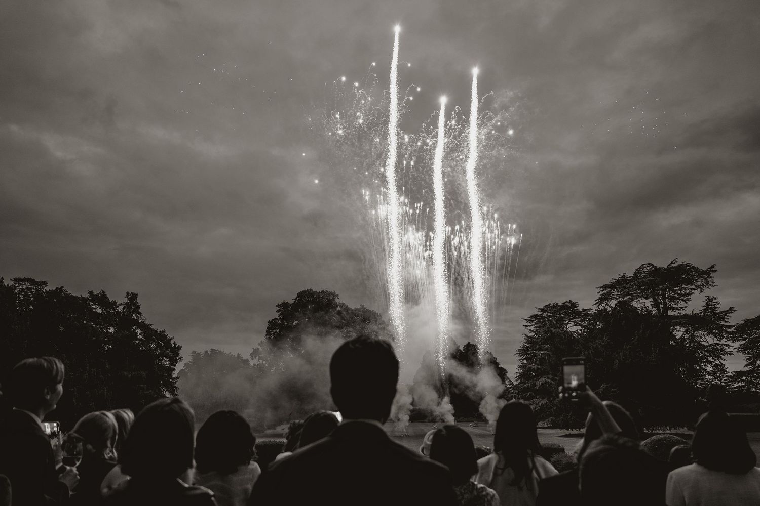 A crowd of silhouetted spectators watches bright fireworks streak into a dark cloudy sky during an outdoor display.