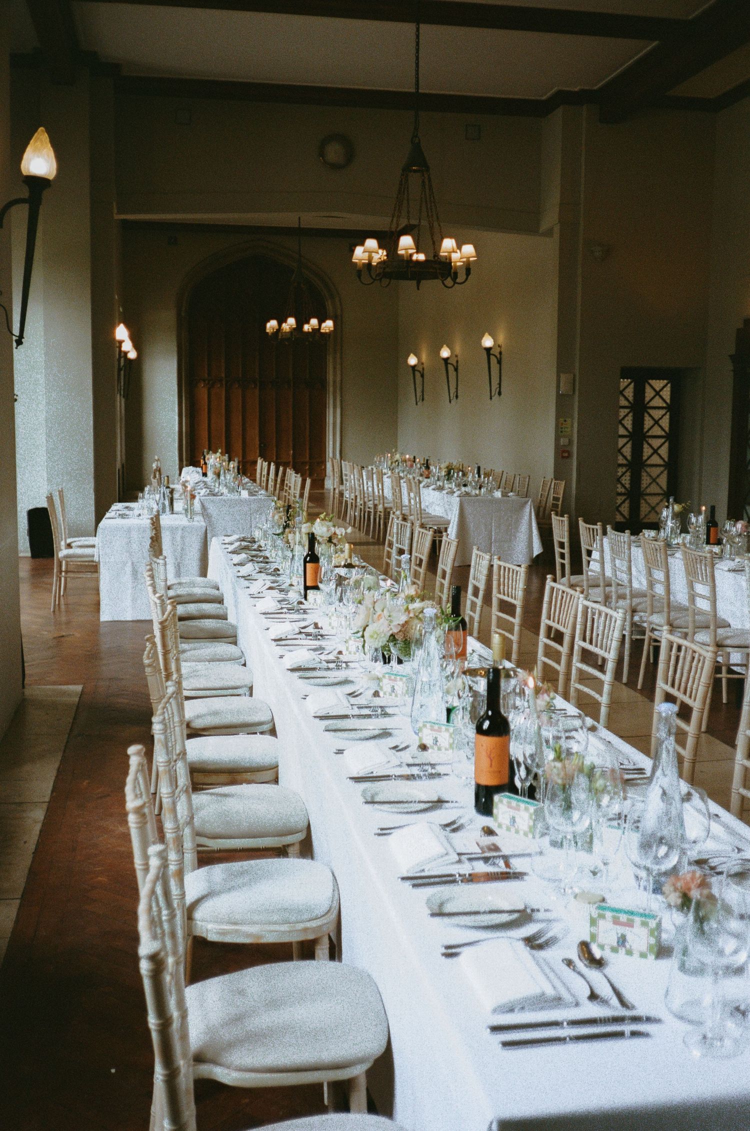 Long formal dining table with white linens and place settings in a dimly lit banquet room with chandeliers.