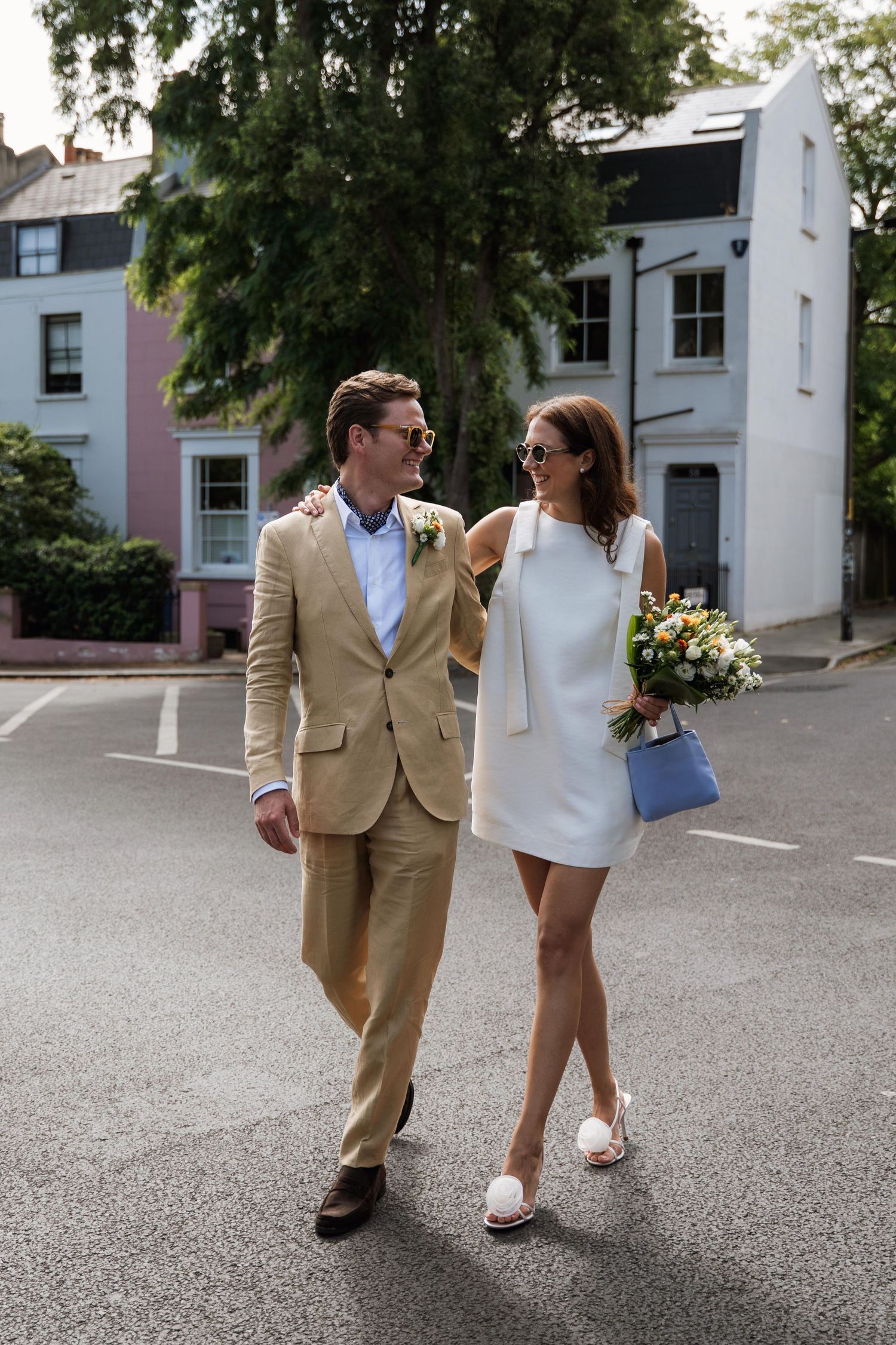 A couple in wedding attire walking together on a paved surface with buildings in the background.