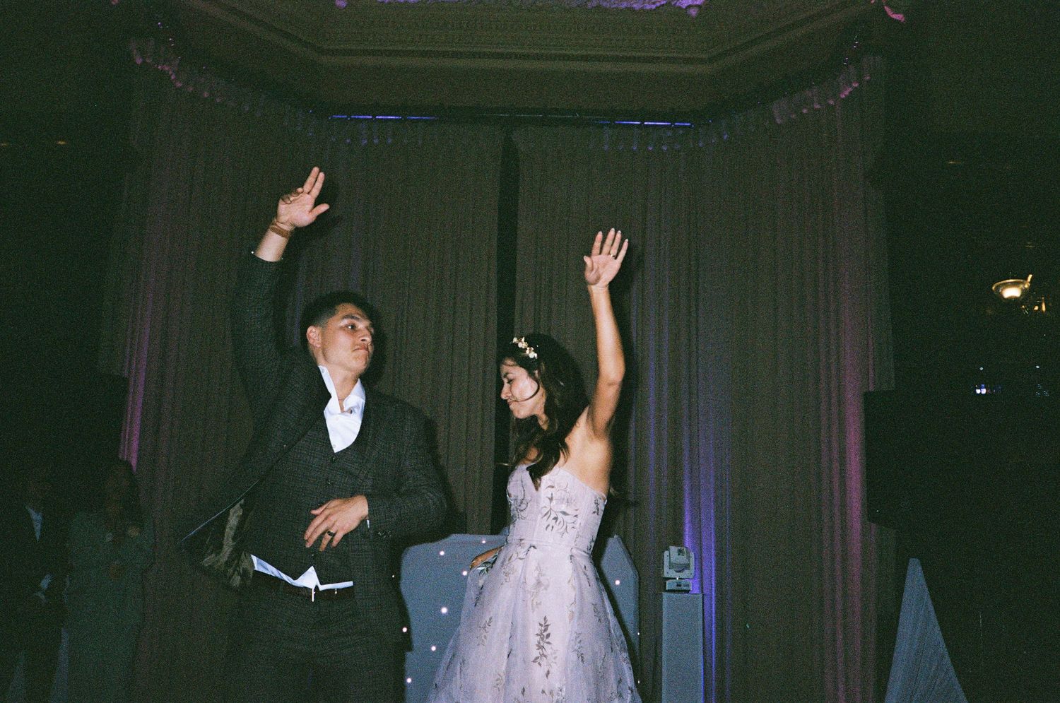 A couple in formal attire joyfully waves to guests at what appears to be a wedding celebration.