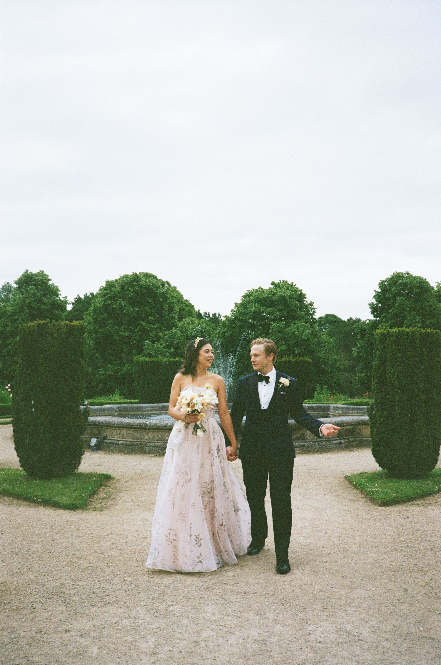A couple walks along a garden path with manicured hedges and fountain in the background on their wedding day.