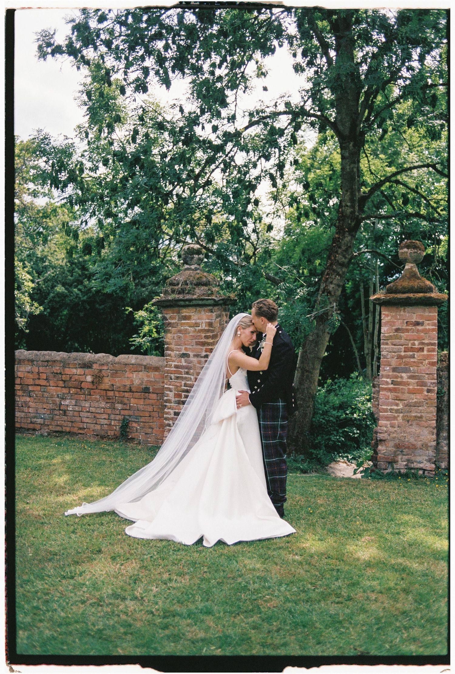 A newlywed couple shares a romantic moment in a garden setting with brick walls and lush greenery.
