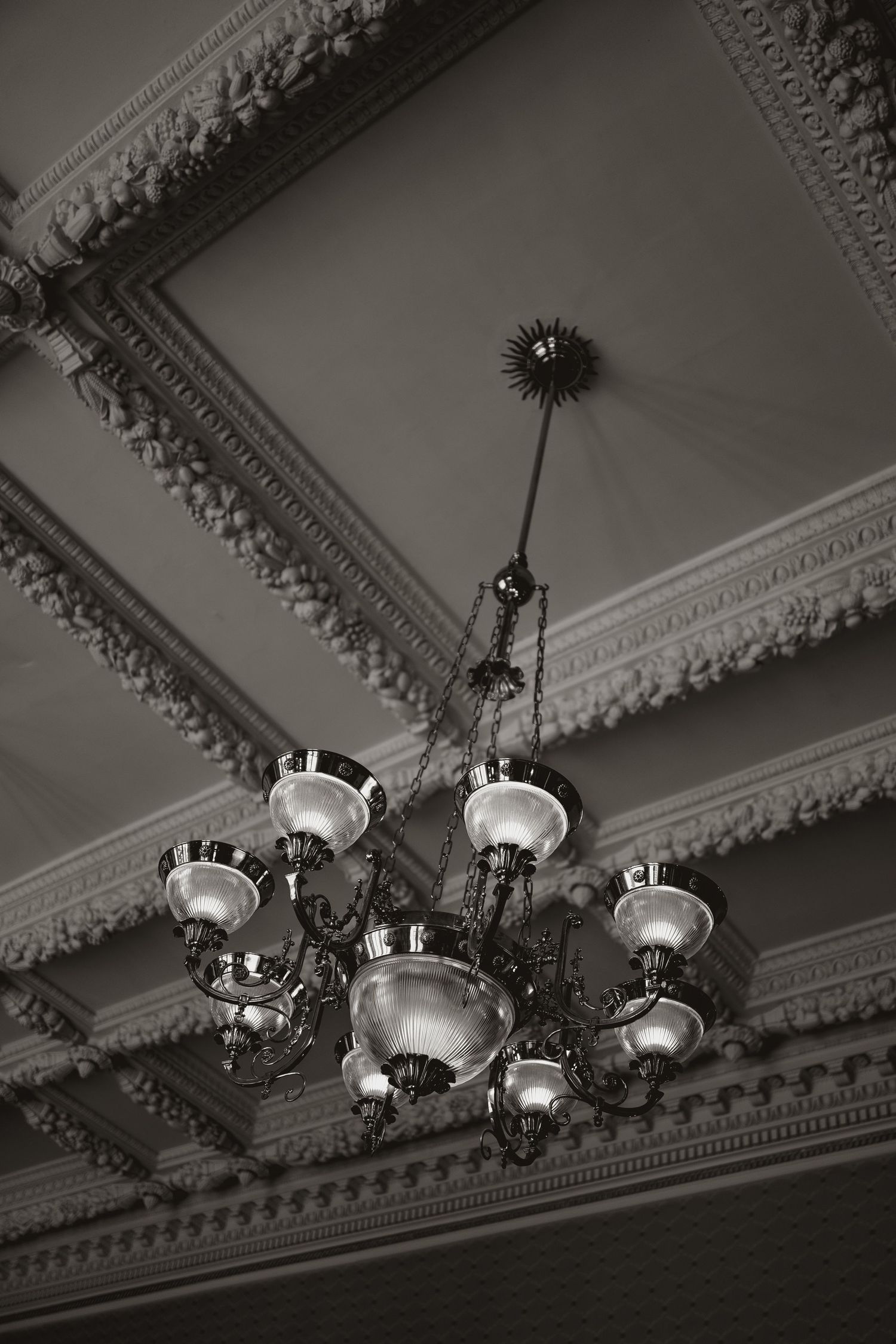 Black and white view of an ornate vintage chandelier hanging from a decorated ceiling with intricate moldings.