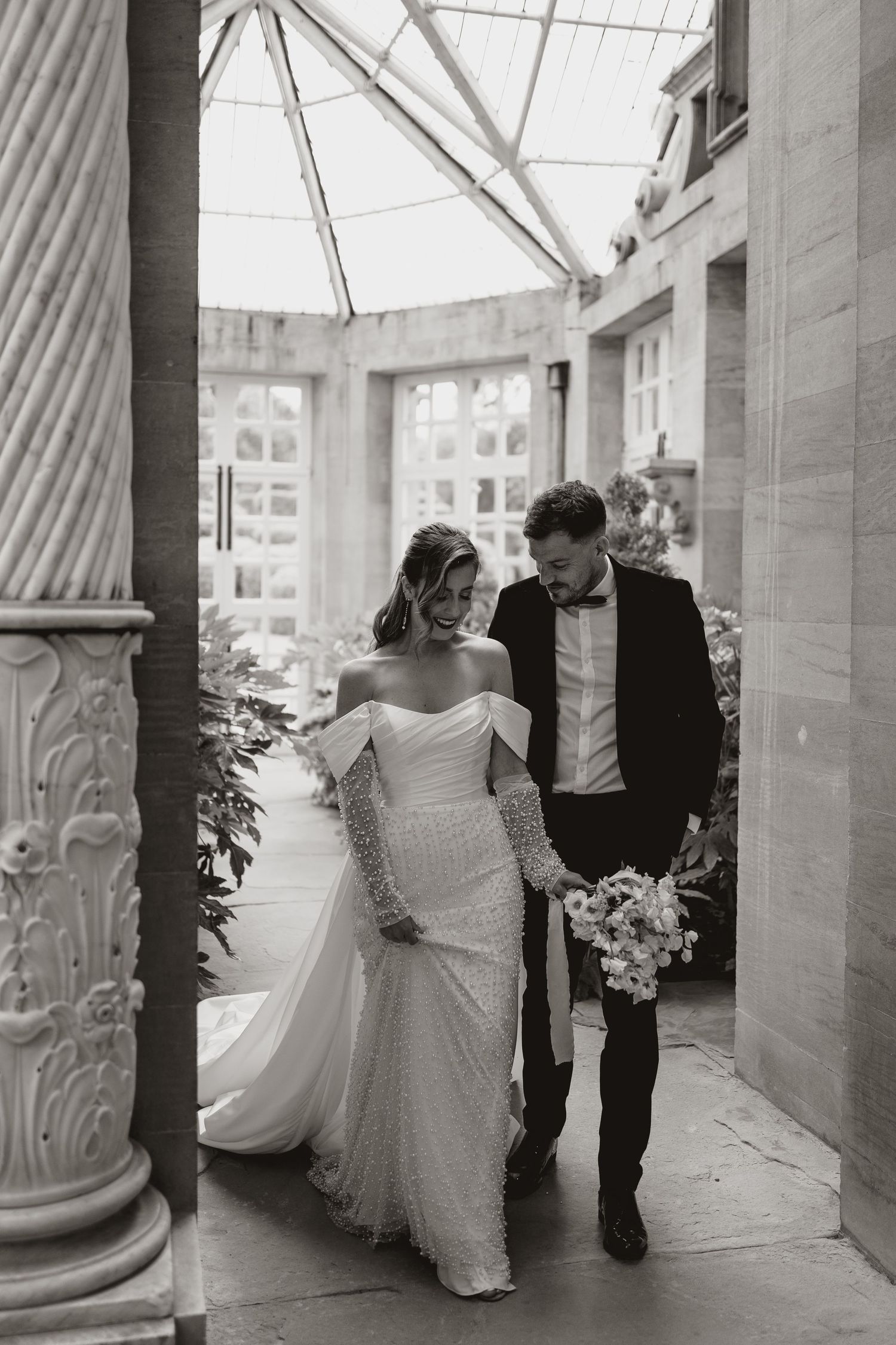 A black and white wedding photograph of a couple walking together in an elegant columned hallway with natural light.