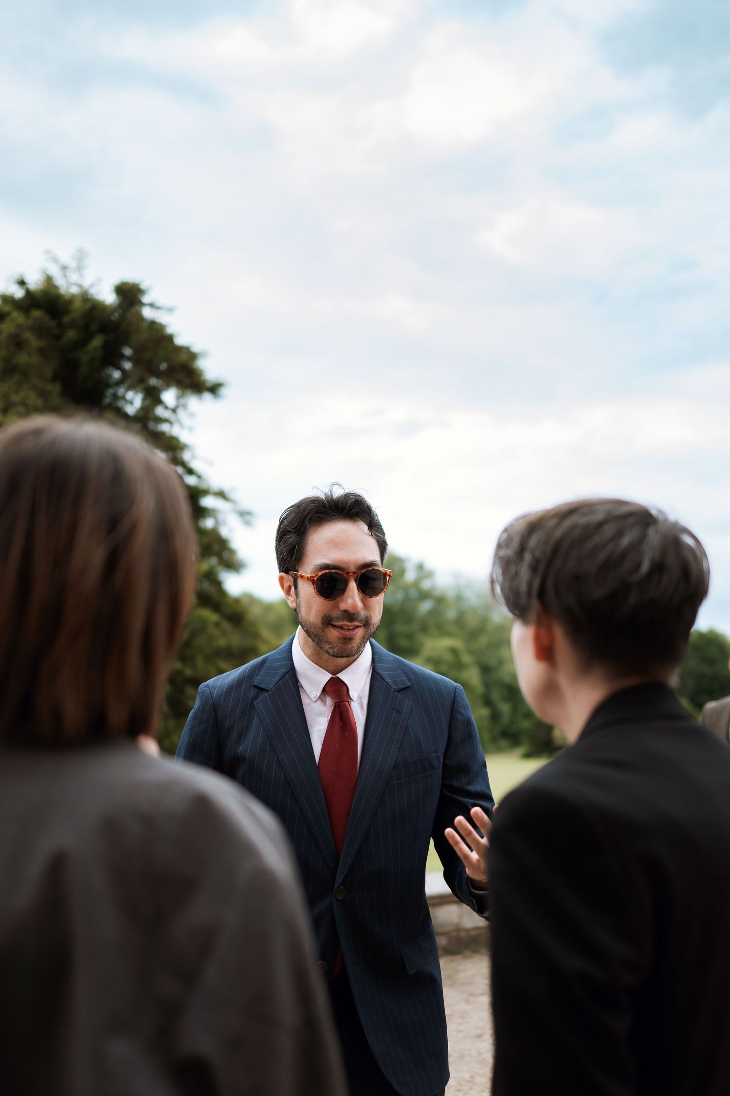 Professional in a navy suit and red tie speaks with colleagues outdoors on a cloudy day.