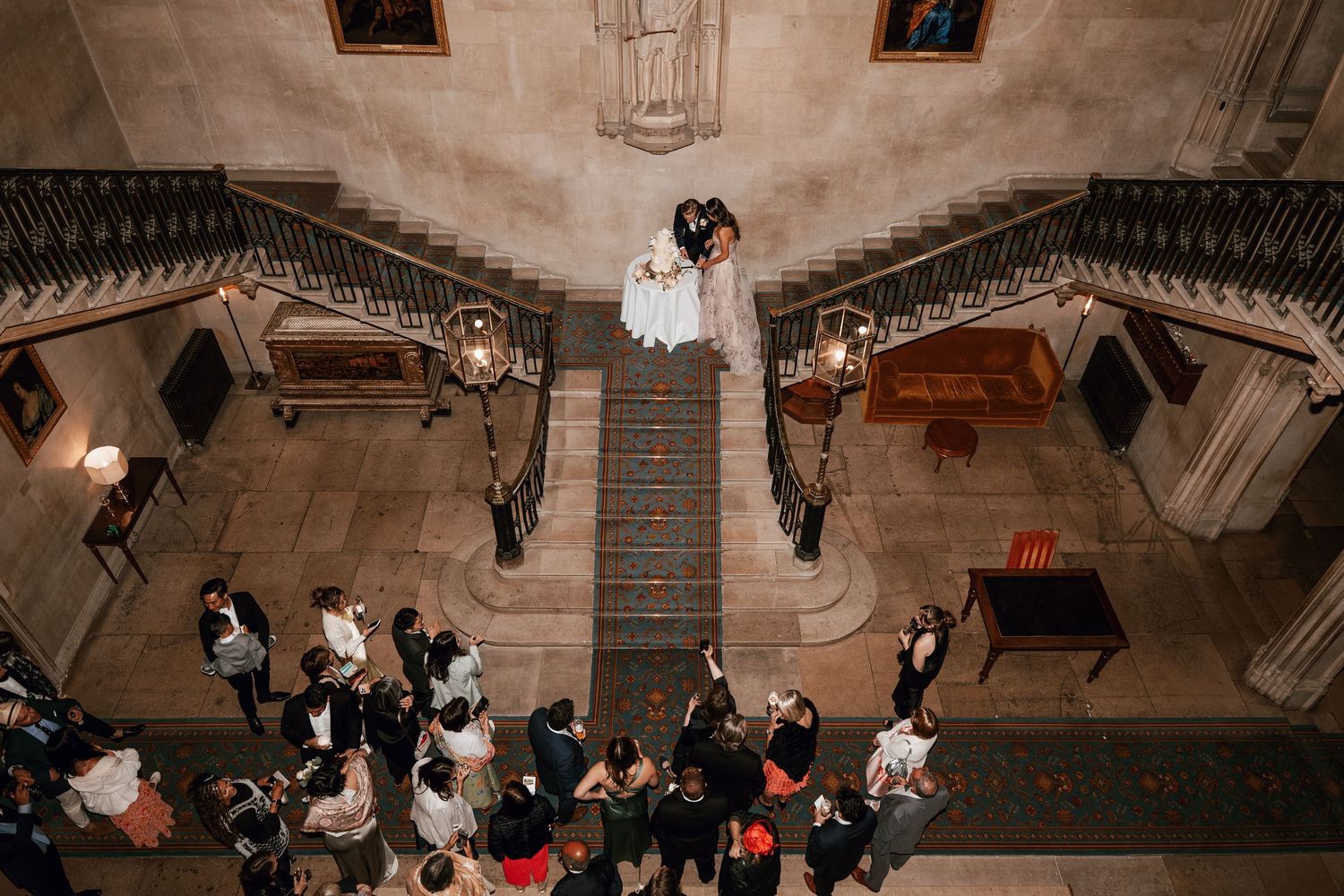 A bird's eye view of a couple on elegant stone staircase with guests gathered below in grand historic venue.