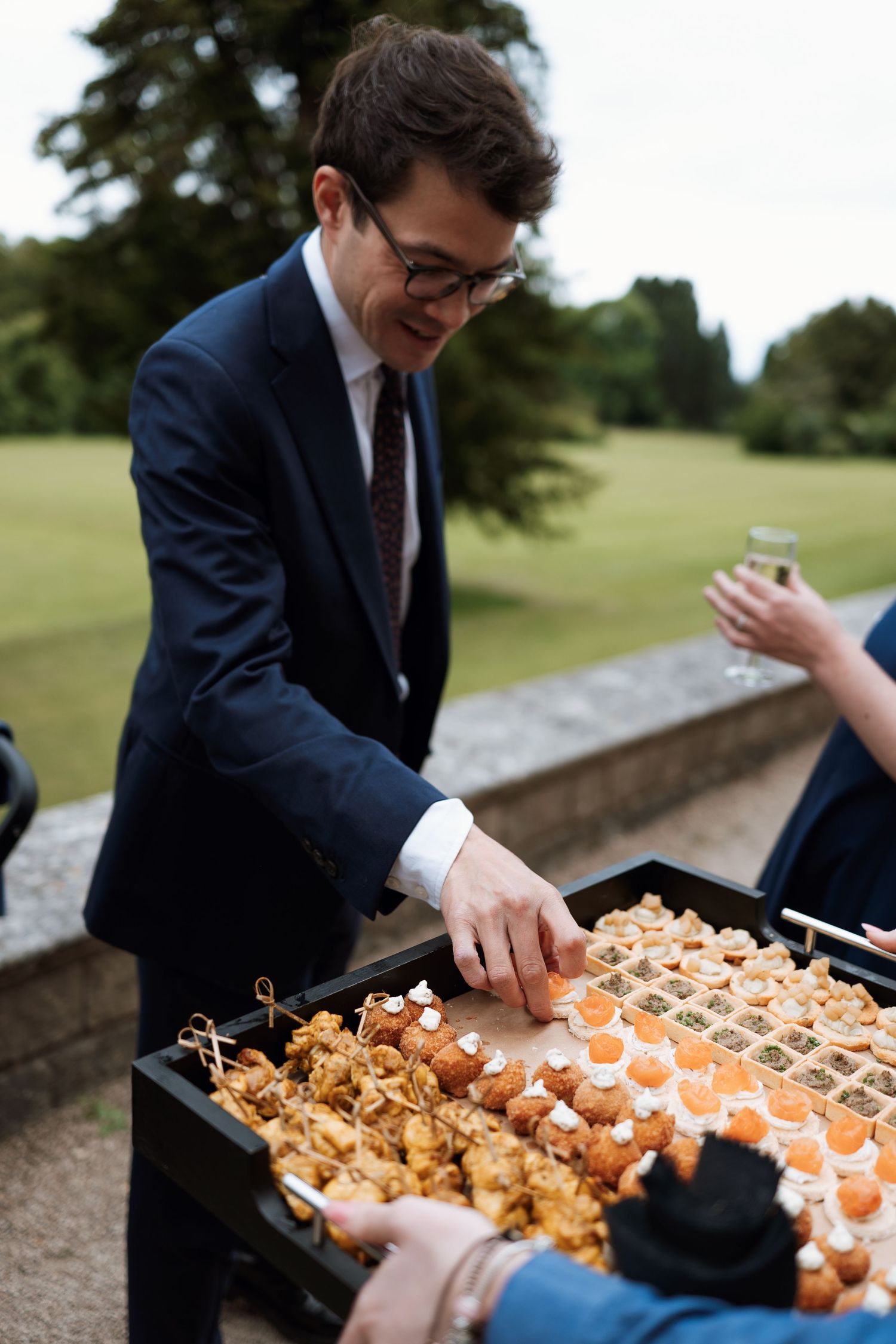Someone in a navy suit selecting appetizers from a catering display tray at an outdoor event.