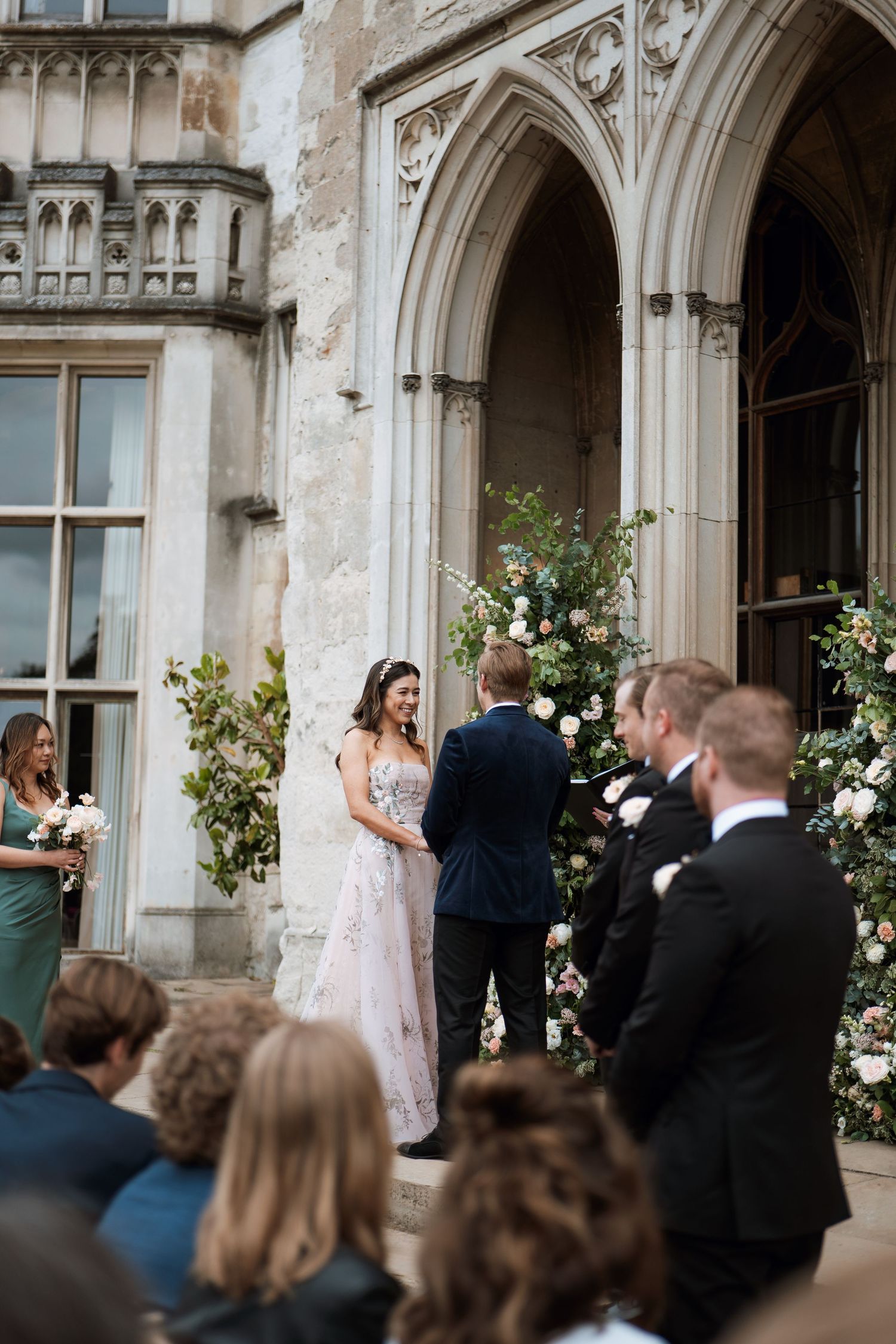 Elegant outdoor wedding ceremony at a Gothic-style venue with ornate stone archways decorated with white floral arrangements.