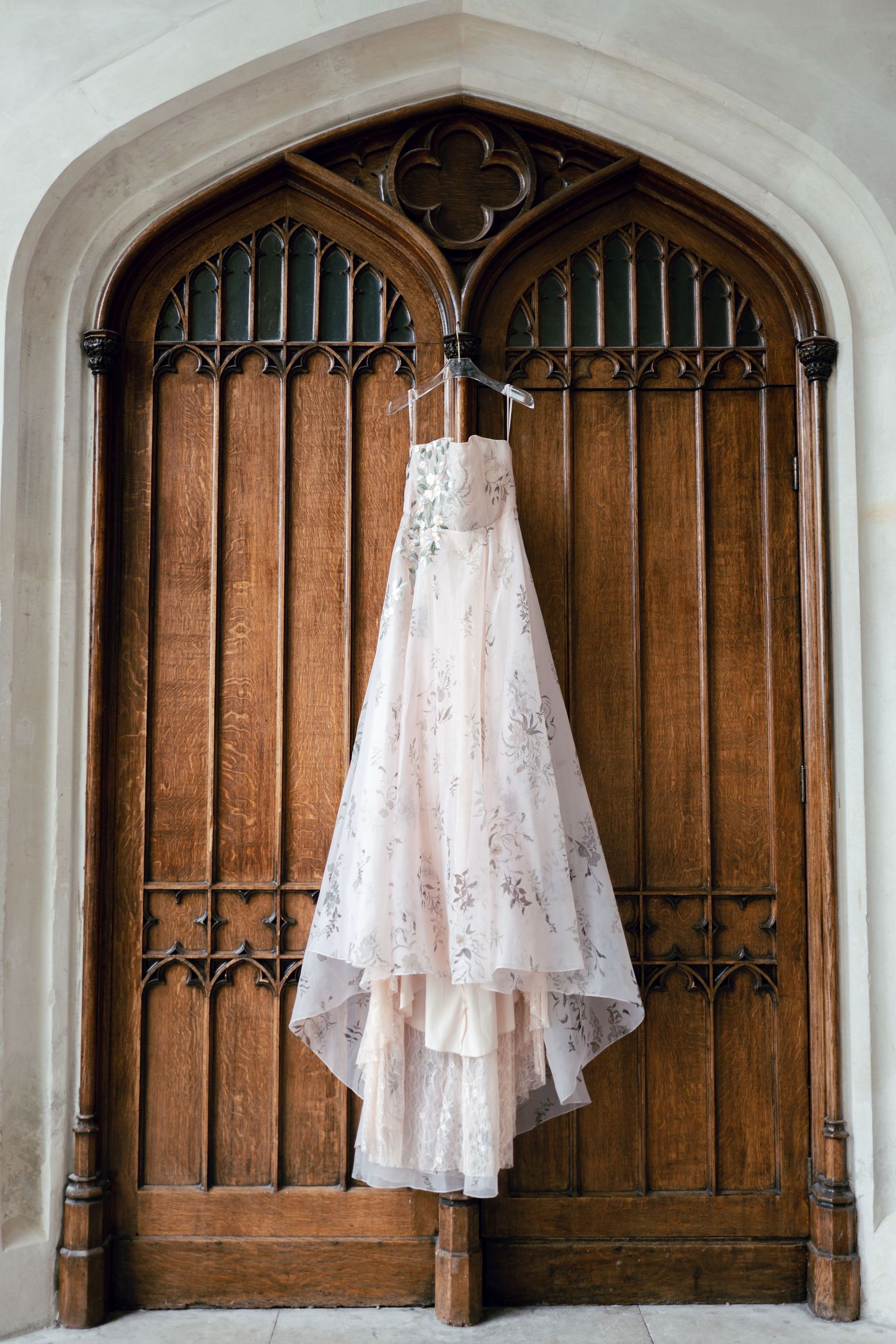 A delicate lace wedding dress hangs on ornate wooden Gothic church doors.