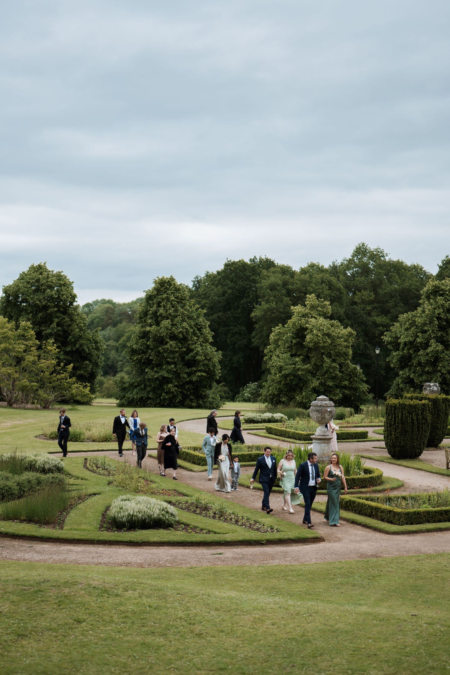 A group of people walking through a formal garden with manicured hedges and pathways under an overcast sky.