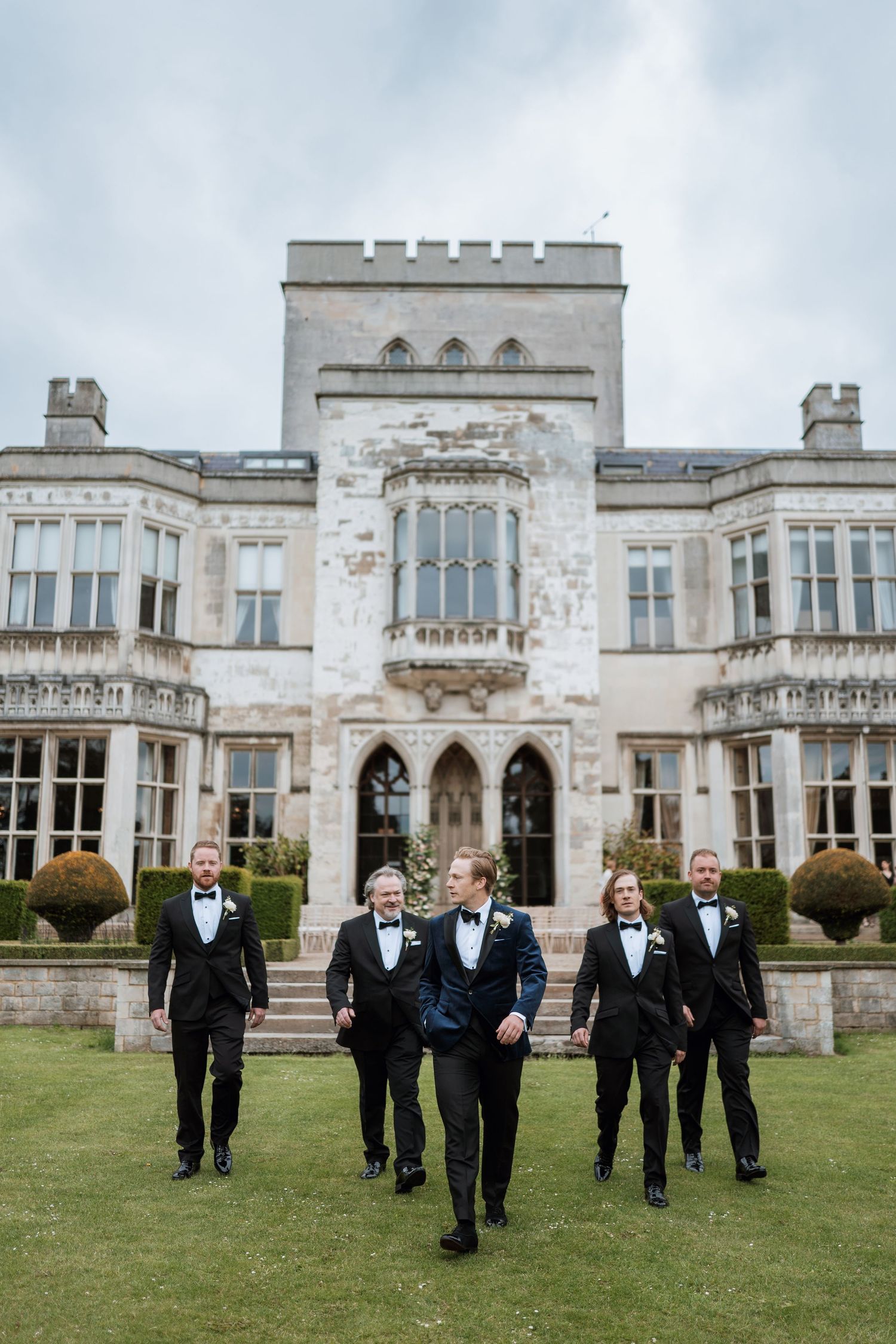 A wedding party group walks across the lawn of an elegant Gothic revival mansion with ornate architecture.