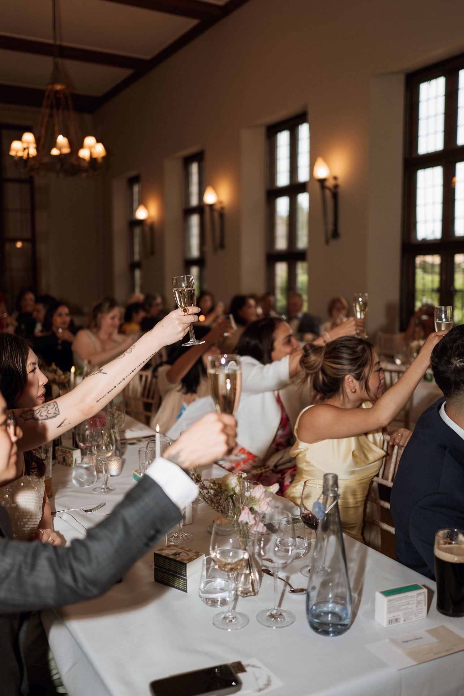 Wedding reception guests raise their glasses in a toast at an elegant indoor venue with tall windows and wall sconces.