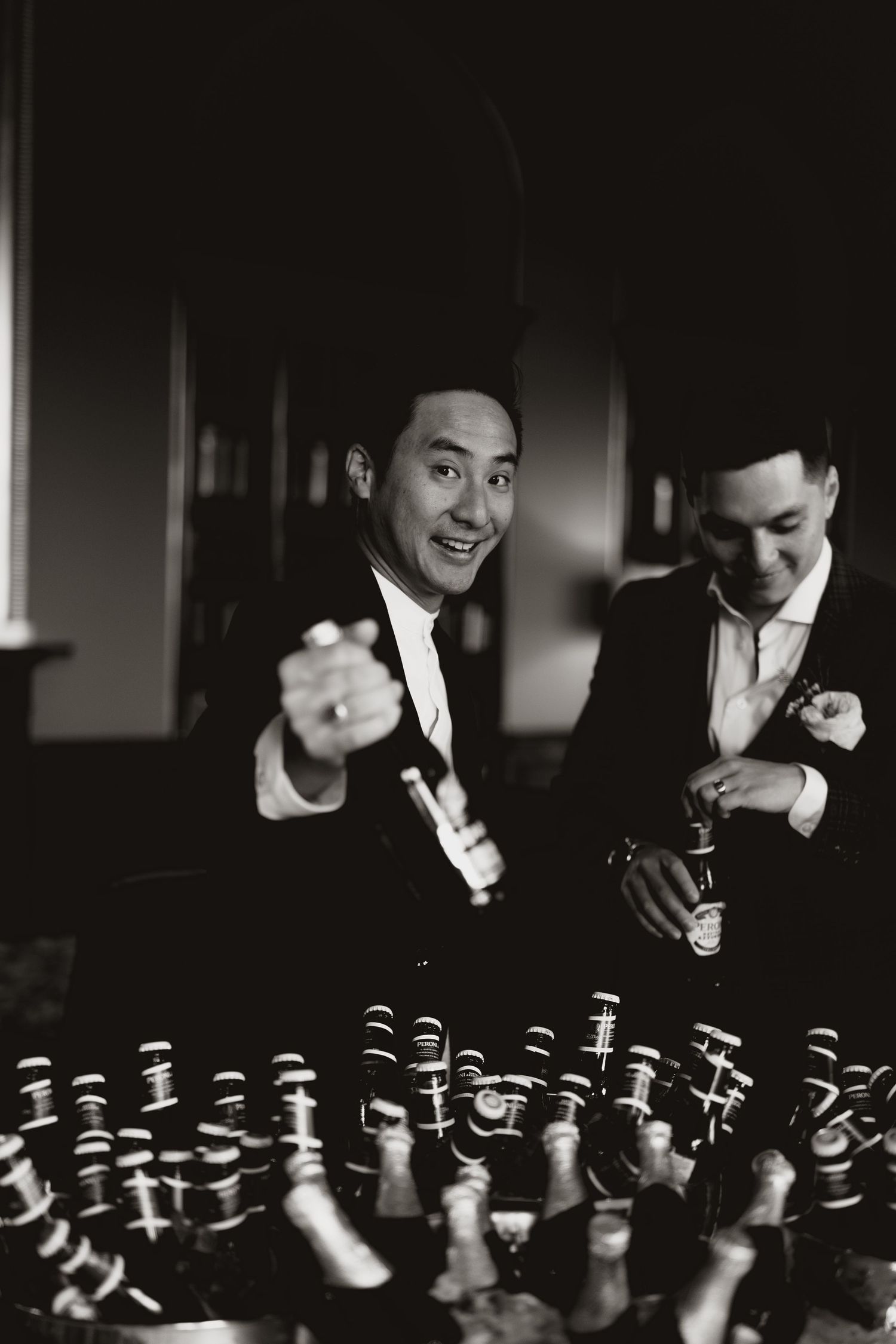 Two men in formal suits celebrate and smile at a party with many bottles displayed on a table in black and white.