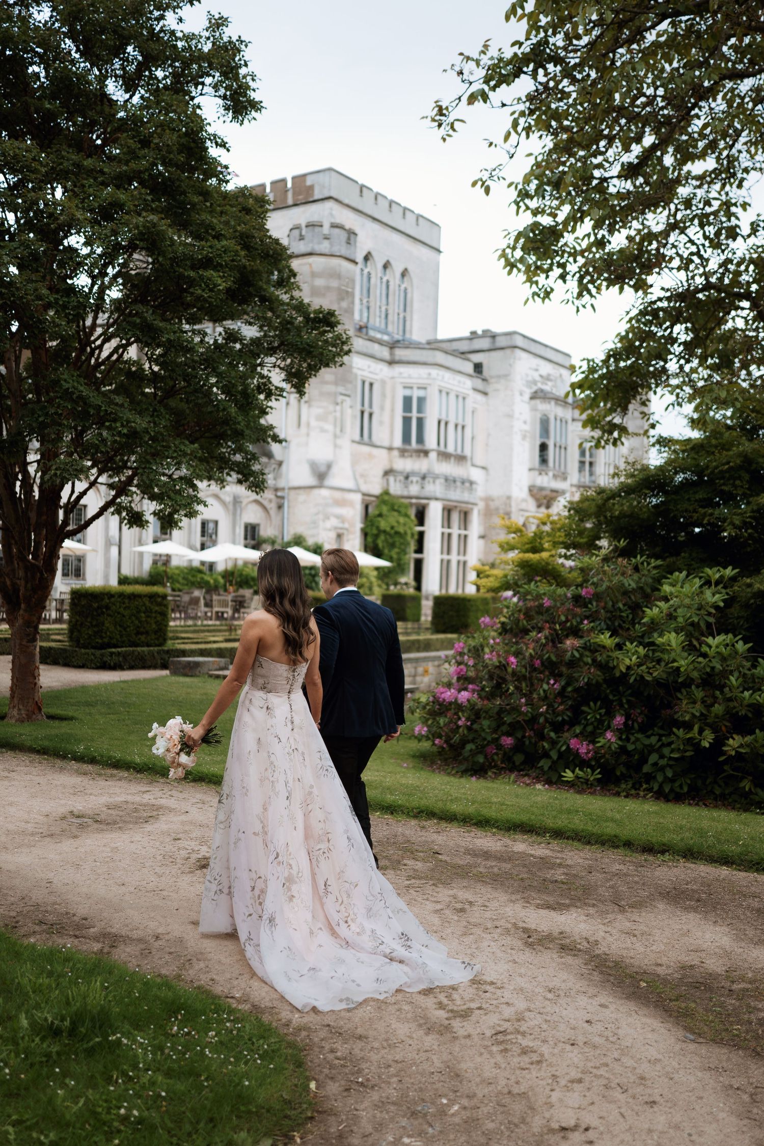 Newlywed couple walks along garden path with their dog beside a grand white Victorian mansion on their wedding day.