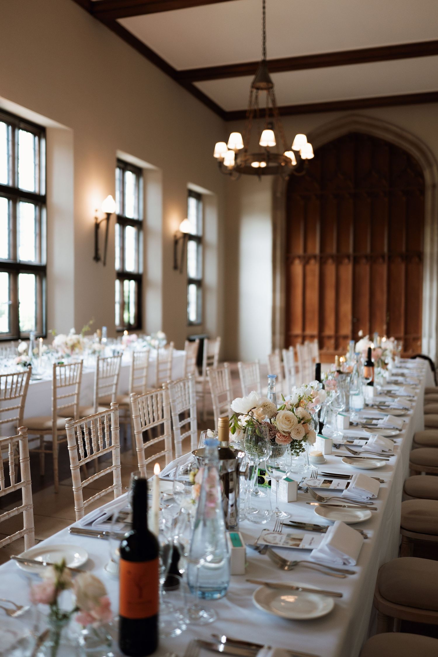 Elegant wedding reception table setting with white linens, floral centerpieces and high windows in a grand dining hall.
