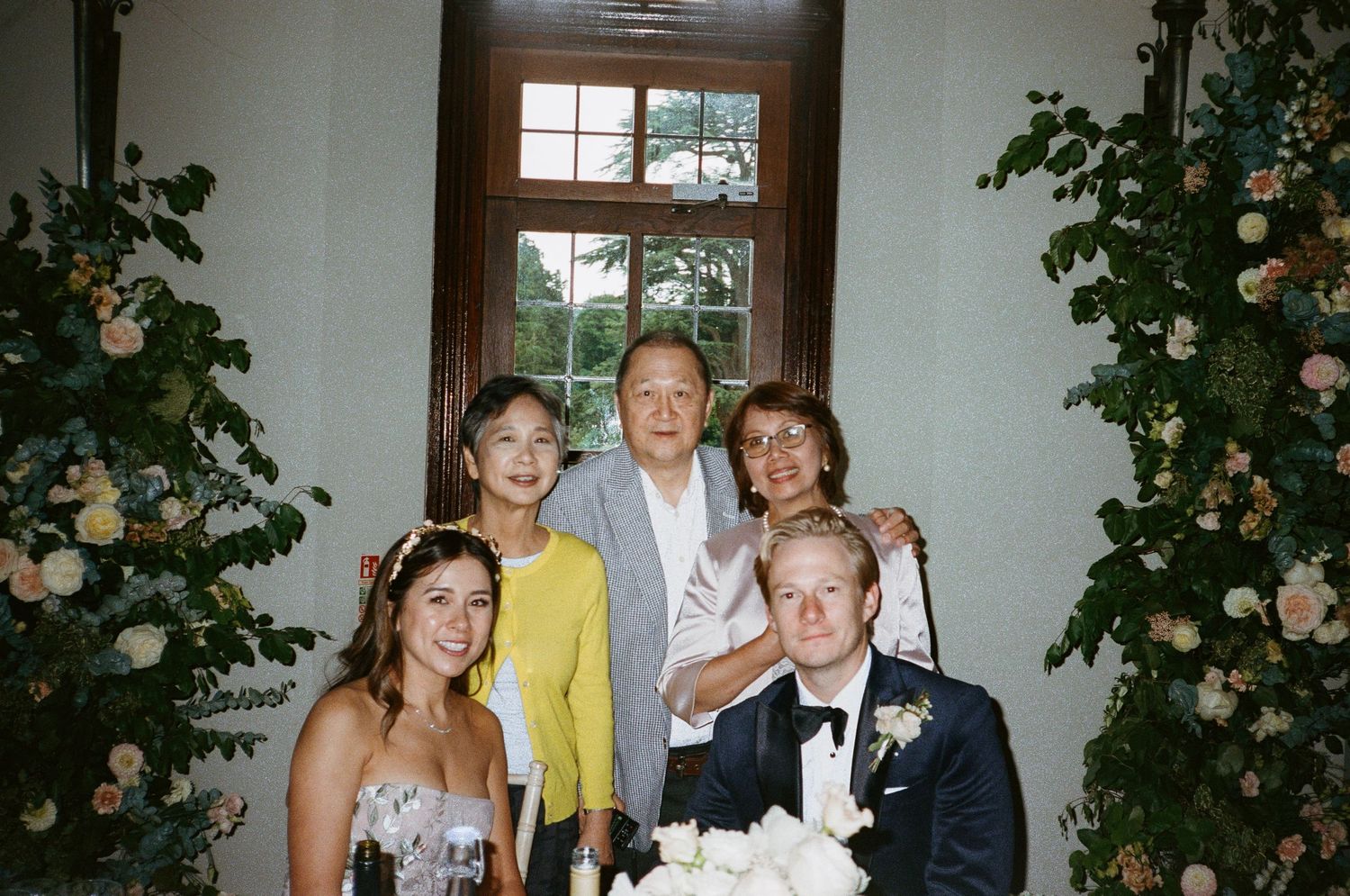 A group stands together at a formal wedding event with floral arrangements and interior decor visible.