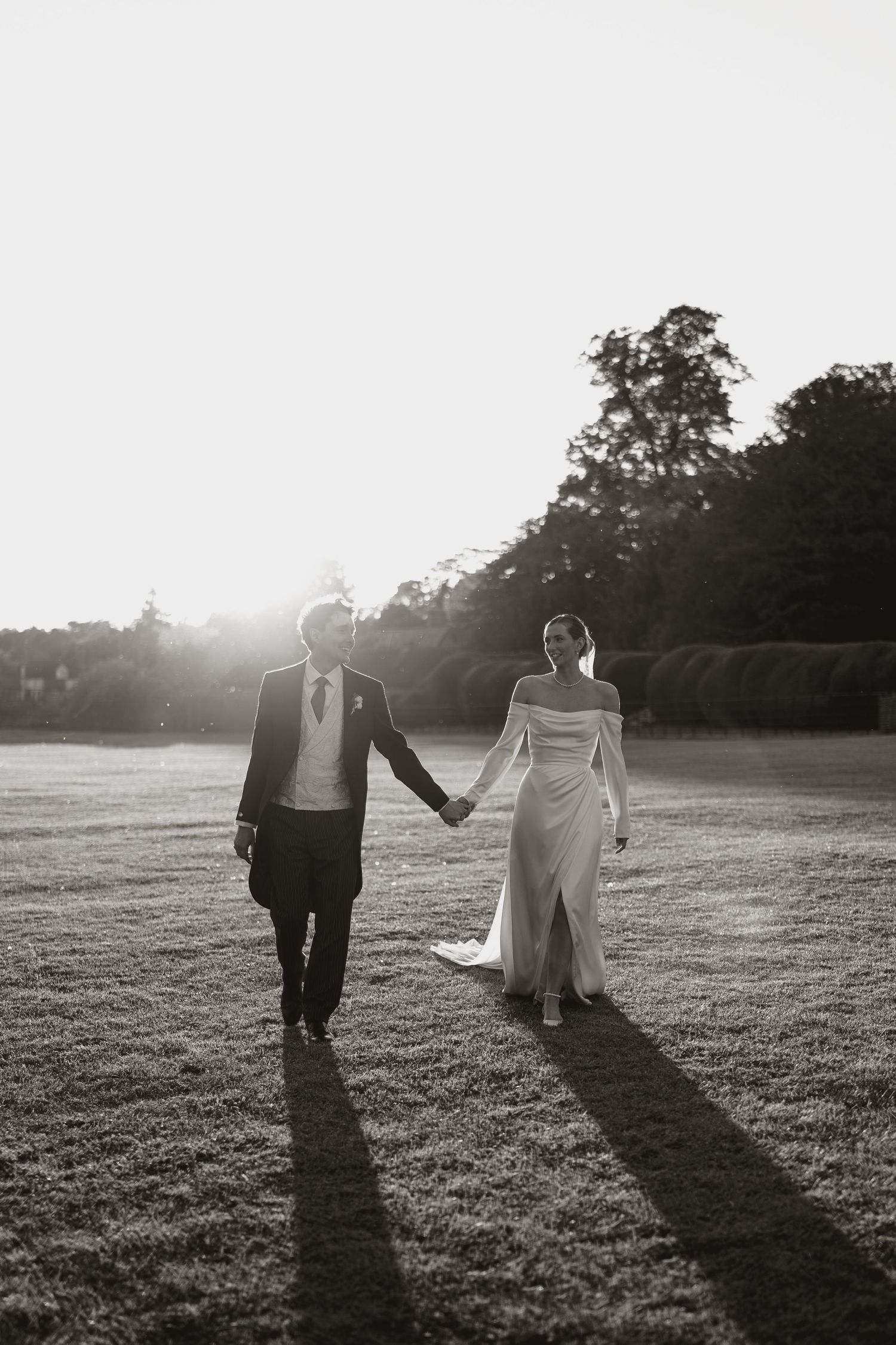 Black and white silhouette of newlyweds walking together in a sunlit field at sunset casting long dramatic shadows.