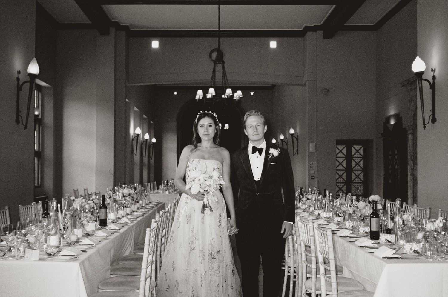 A couple stands in an elegant reception hall with long tables lined with formal place settings and soft lighting.