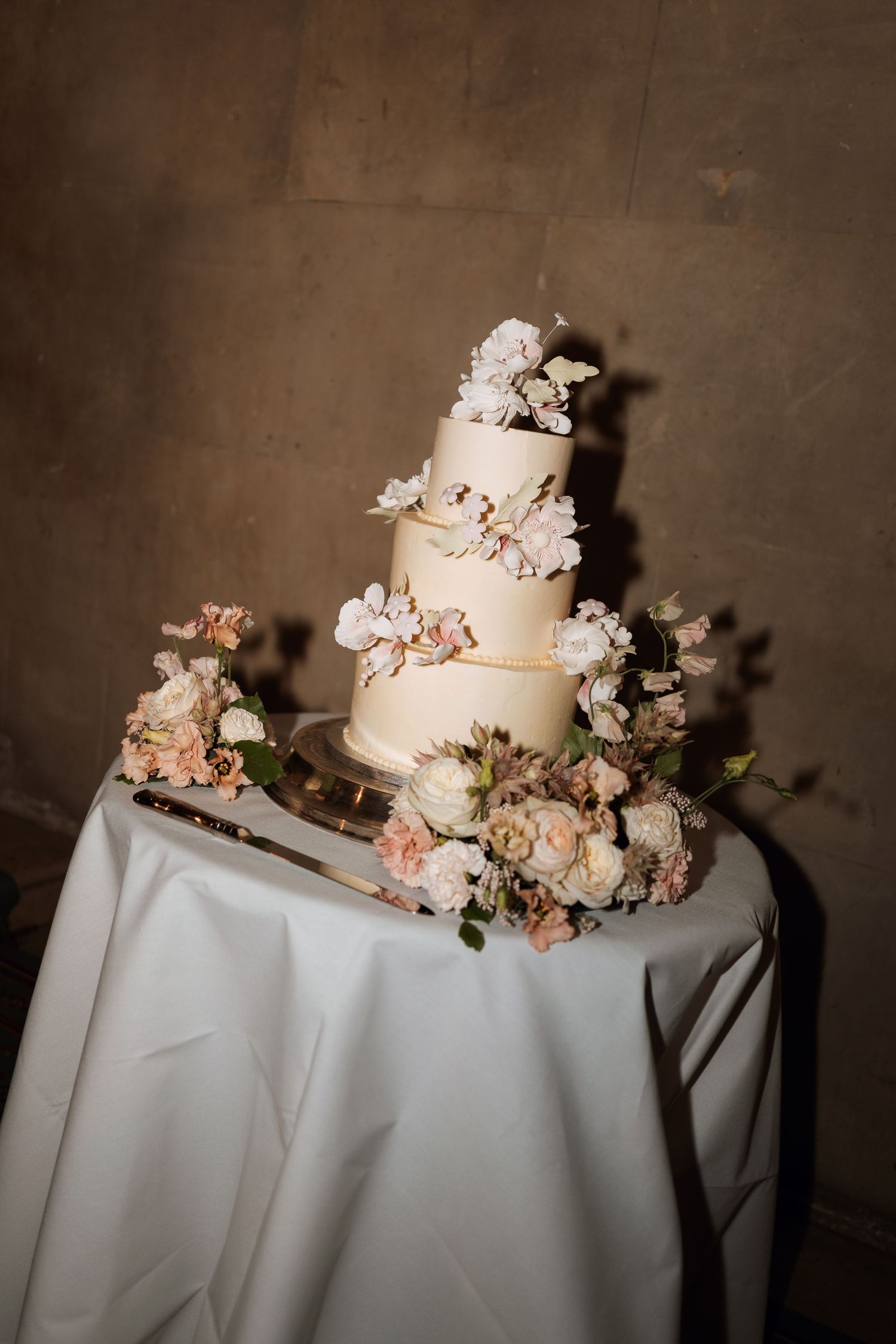 An elegant four-tier white wedding cake decorated with blush pink flowers and greenery sits on a white tablecloth.