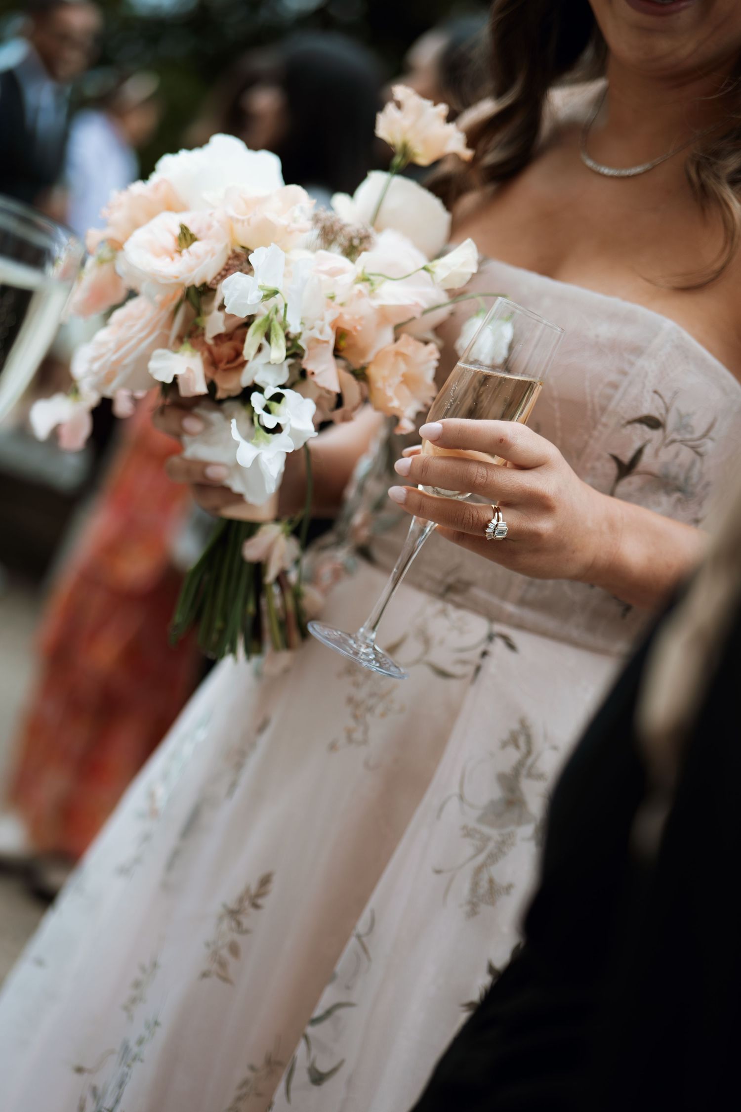 Person in embroidered wedding dress holds champagne glass and delicate peach and white floral bouquet.