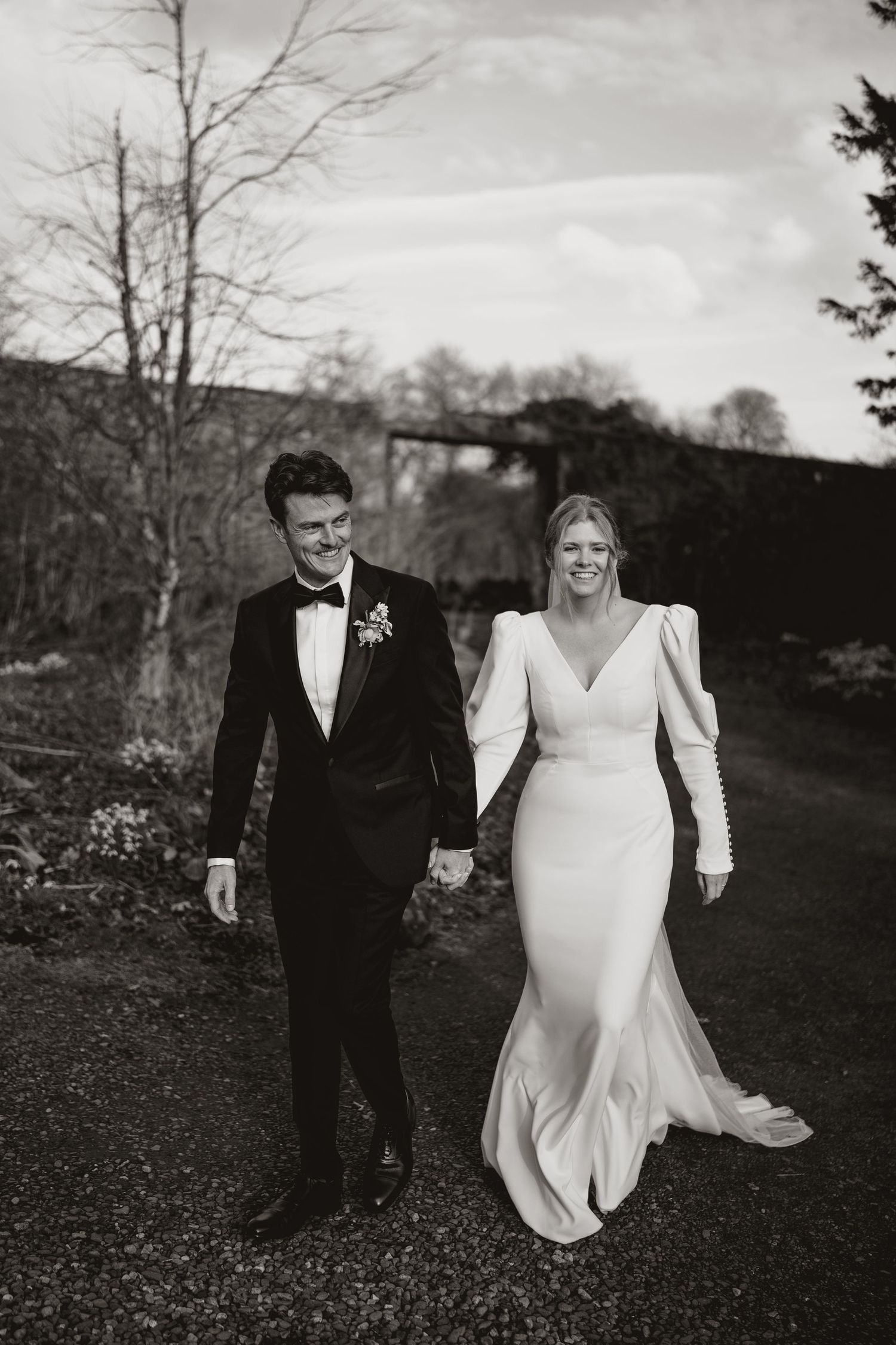 A couple walking together in formal wedding attire on a gravel path in a black and white photograph.