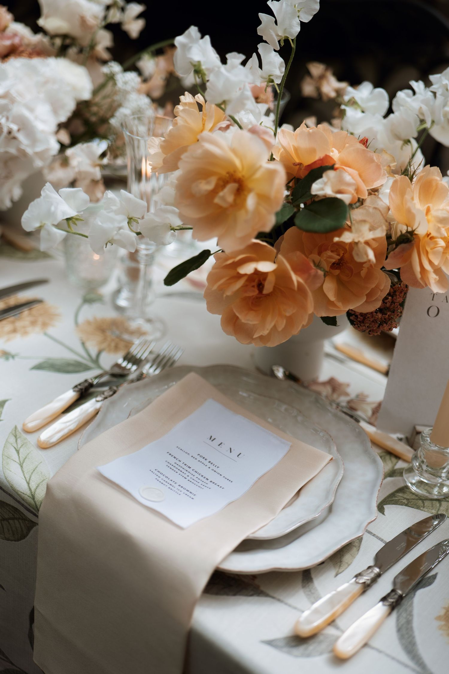 Elegant table setting with peach roses, white flowers, and menu card on cream linens with crystal glassware.