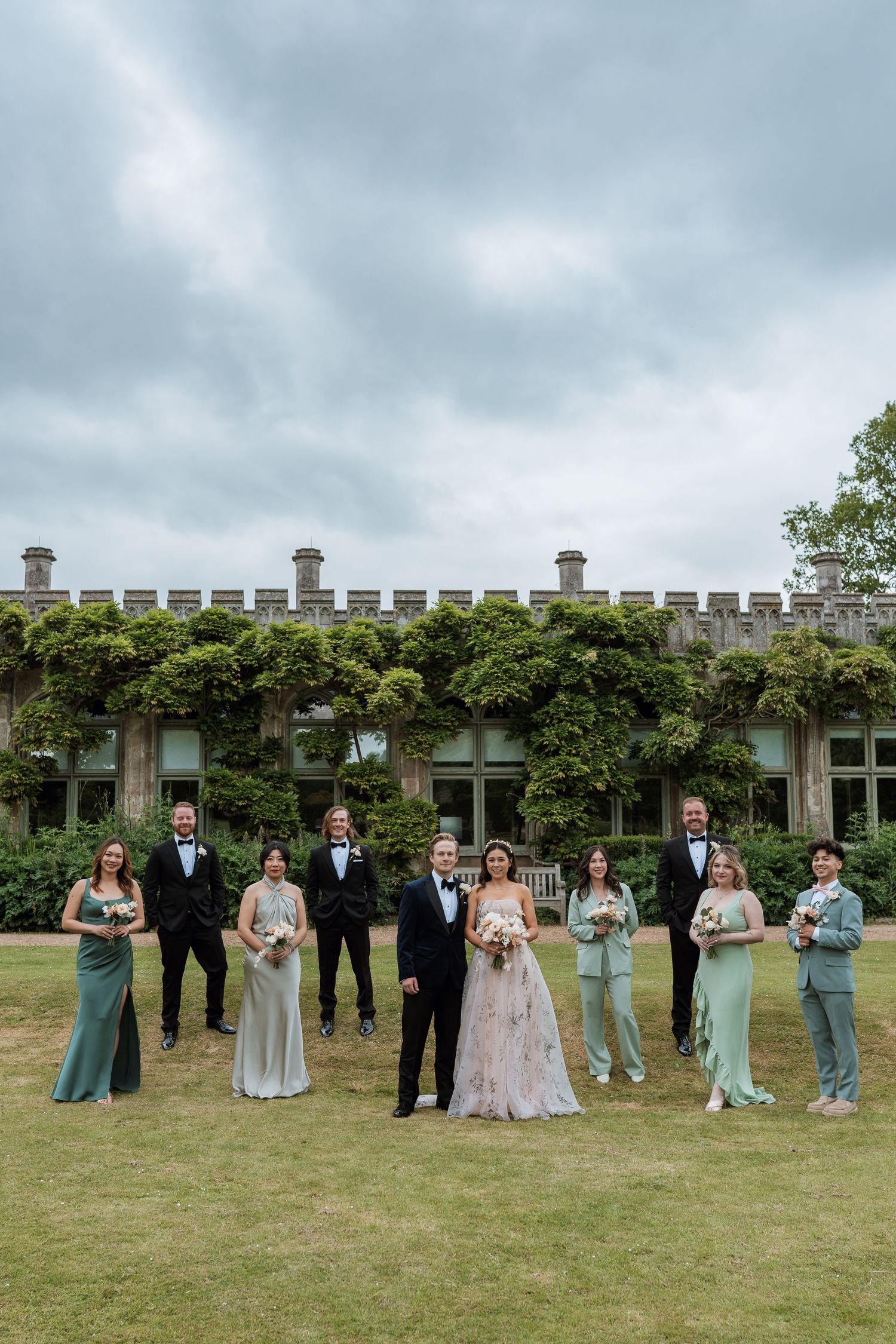 Wedding party poses on lawn in front of ivy-covered stone building on overcast day.
