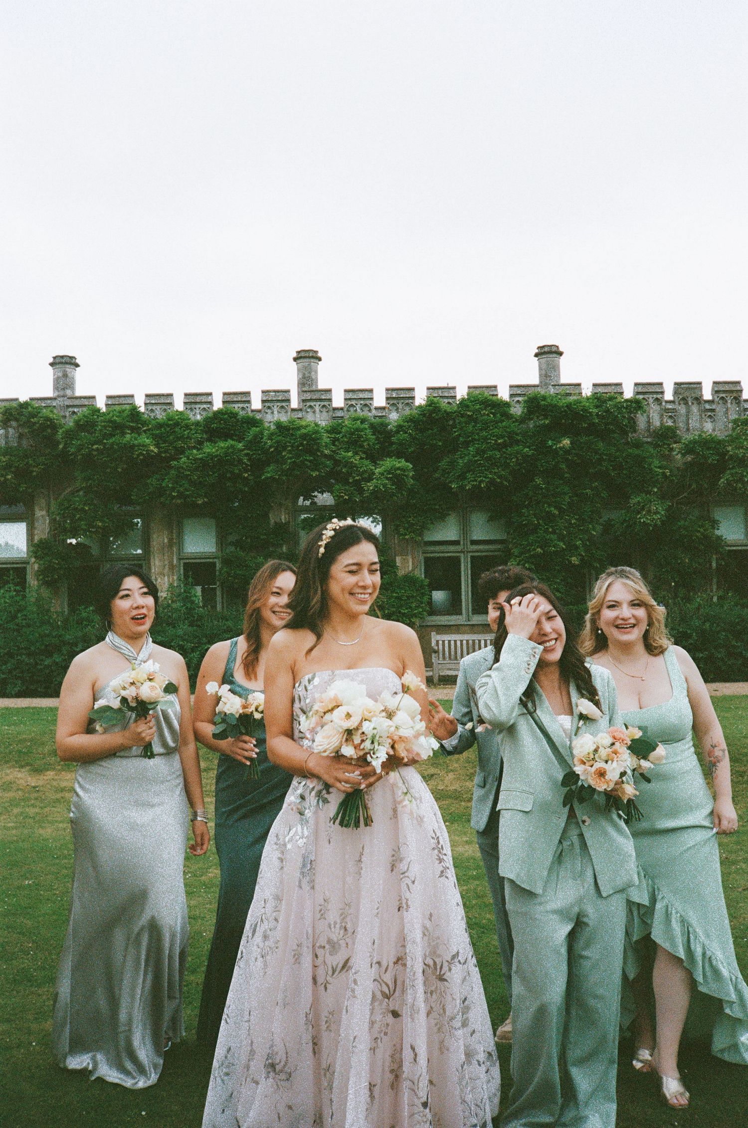 Wedding party poses outdoors with floral bouquets in mint green and pastel dresses against an ivy-covered building.