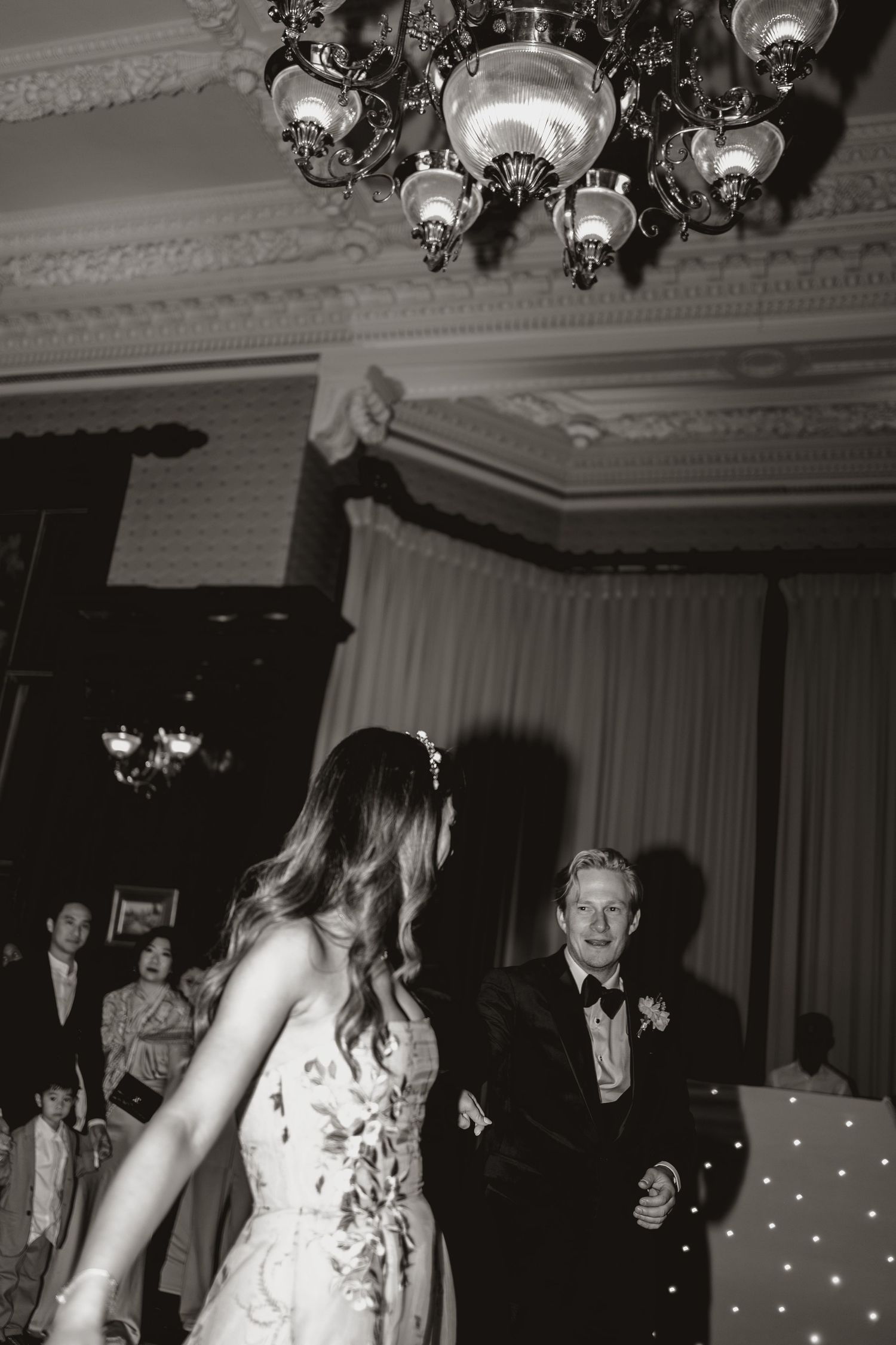 Elegant couple dancing in a formal ballroom under ornate chandelier lighting in a black and white photograph.