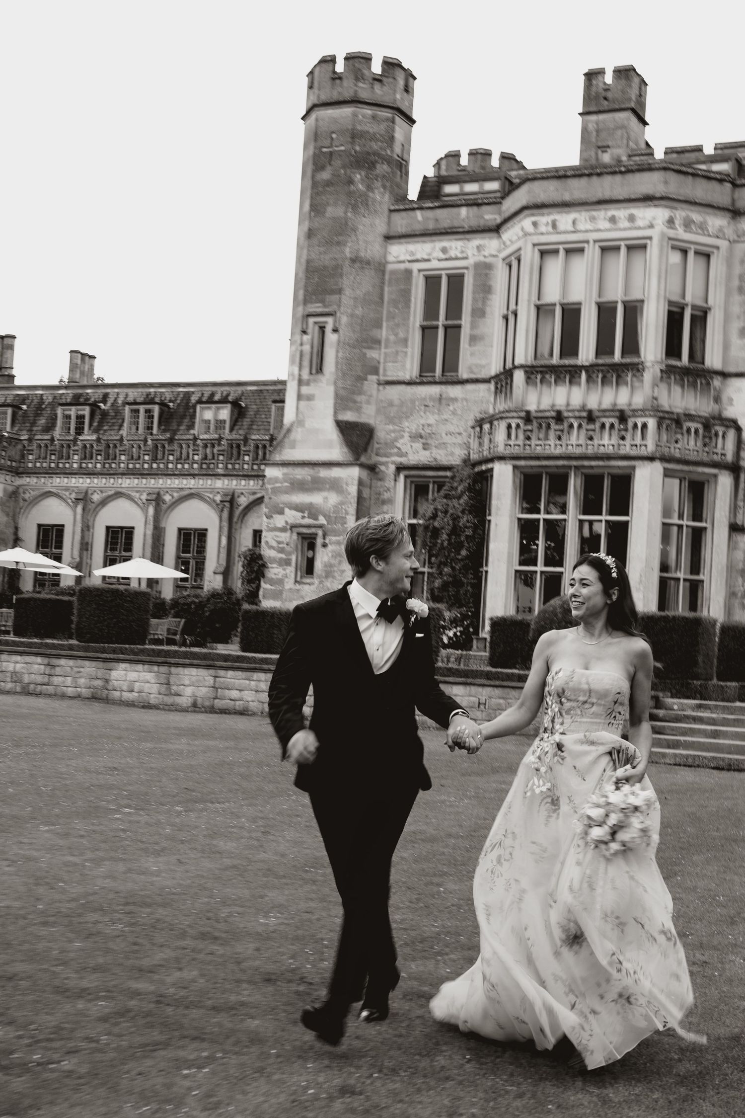 A couple in wedding attire walk hand-in-hand across the lawn of an elegant stone castle with turrets and grand windows.