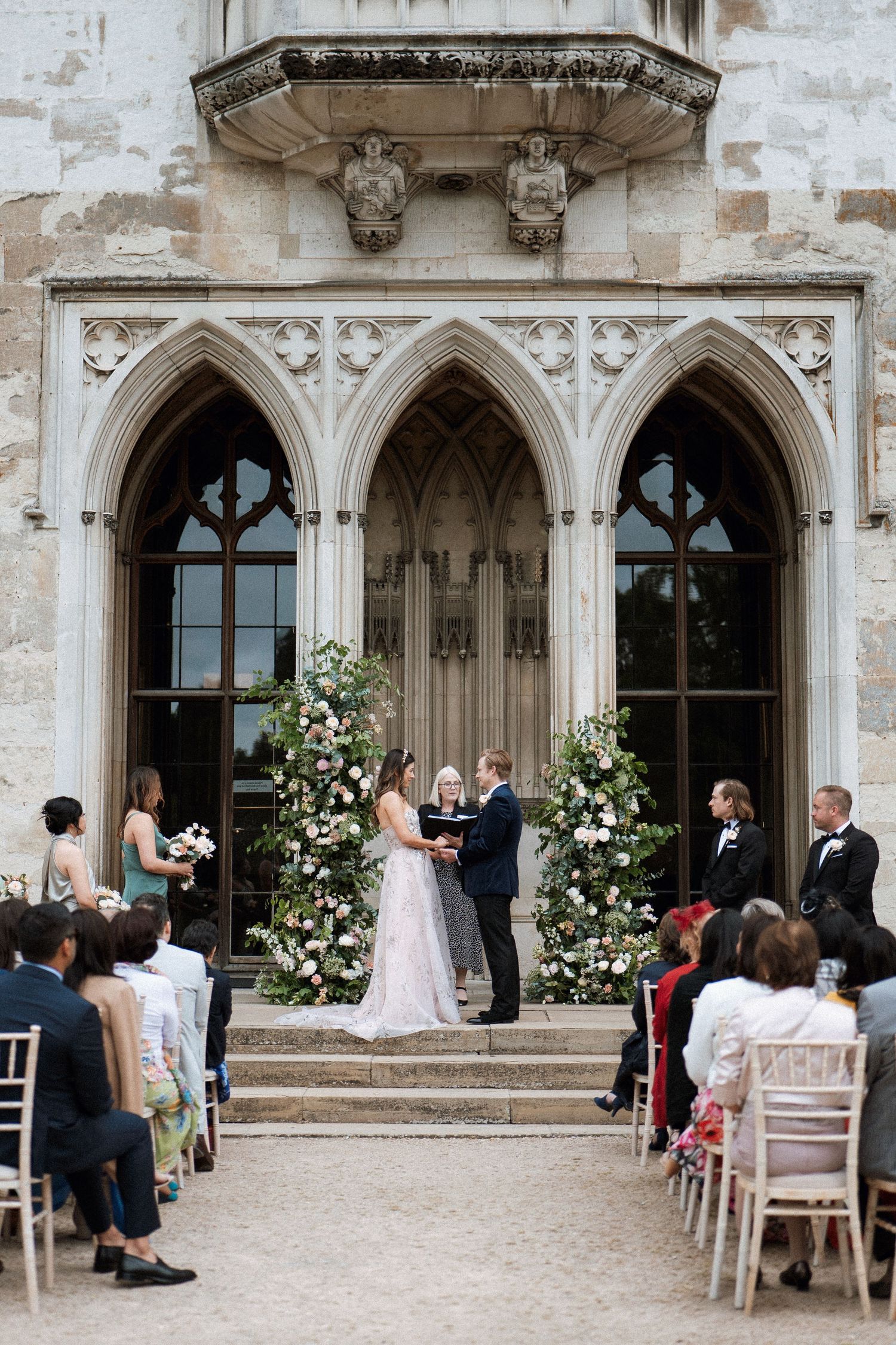 Elegant outdoor wedding ceremony at a gothic-style stone archway decorated with climbing white roses and floral arrangements.