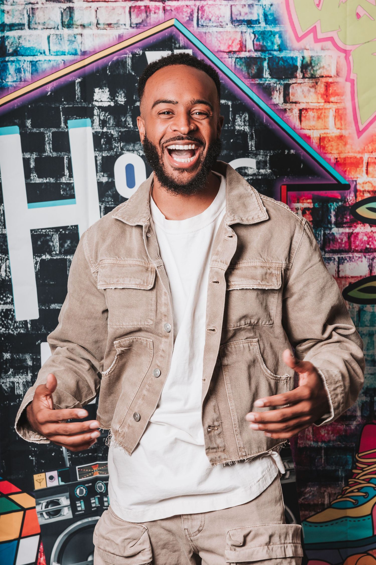 A person wearing a beige jacket and white t-shirt poses with expressive gestures against a colorful graffiti wall background.