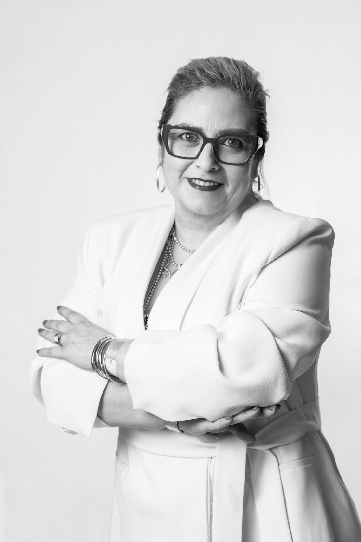 Professional portrait of a person in a white blazer posing with crossed arms, wearing glasses and jewelry against a neutral backdrop.