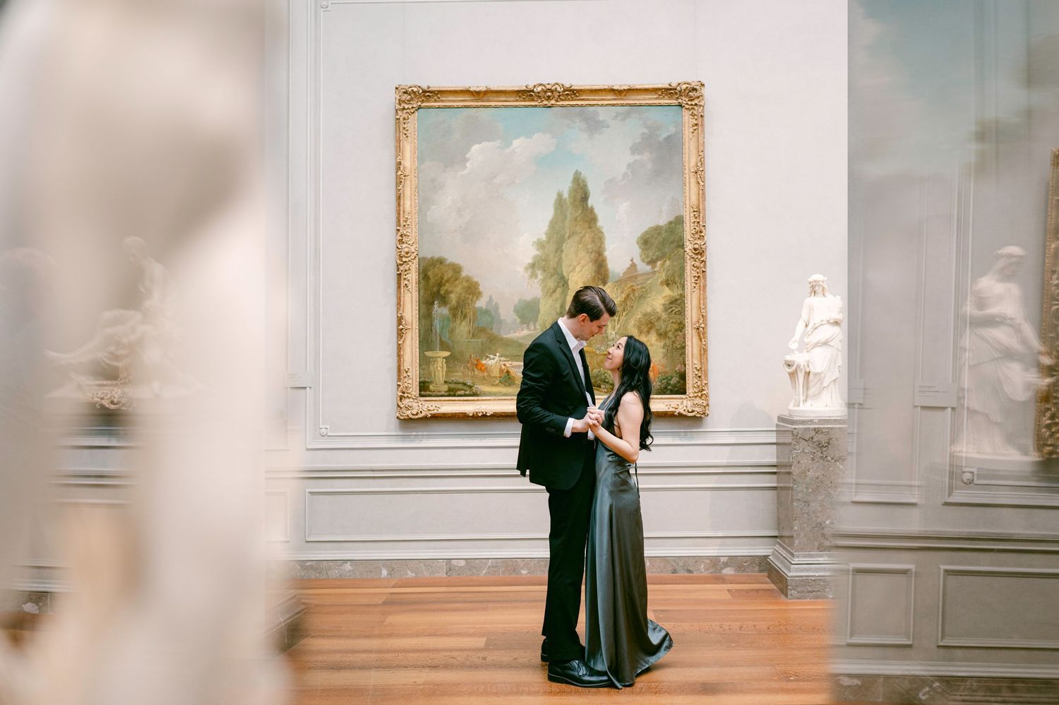 Visitors view classical paintings in an elegant museum gallery with white walls and ornate frames.