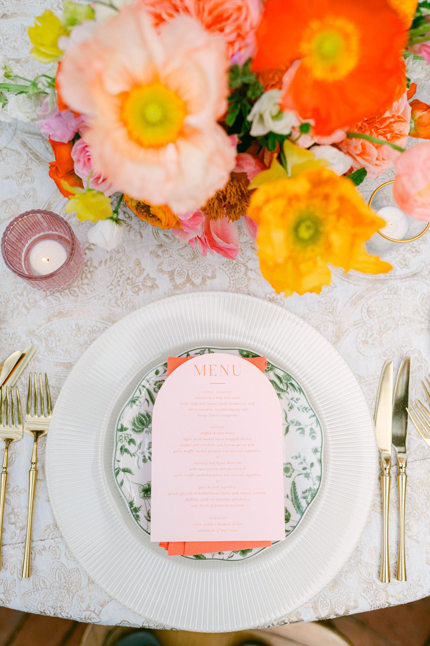 Elegant place setting with pink menu card on floral plate with vibrant orange and yellow poppies in background.