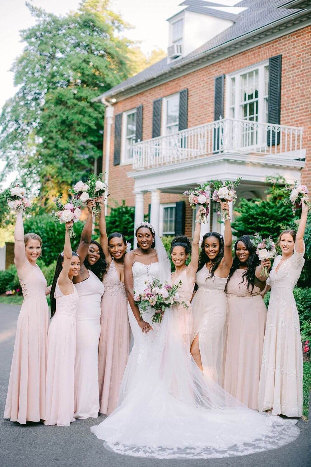 A bridal party in blush pink bridesmaid dresses poses cheerfully outside a brick building at Brittland Manor in Chestertown, Maryland.