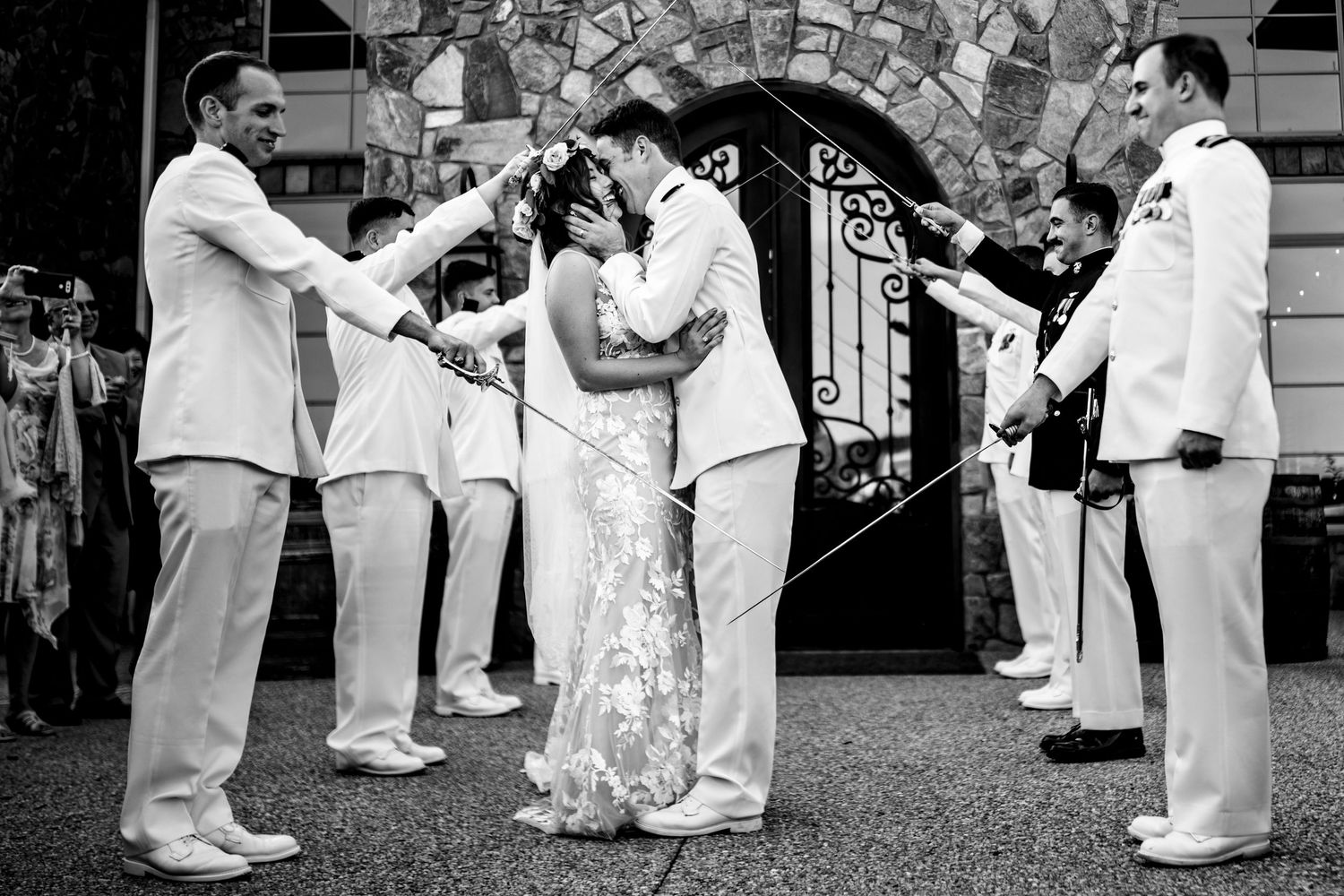 A black and white wedding scene showing a couple sharing a romantic kiss under an arched gateway surrounded by onlookers.