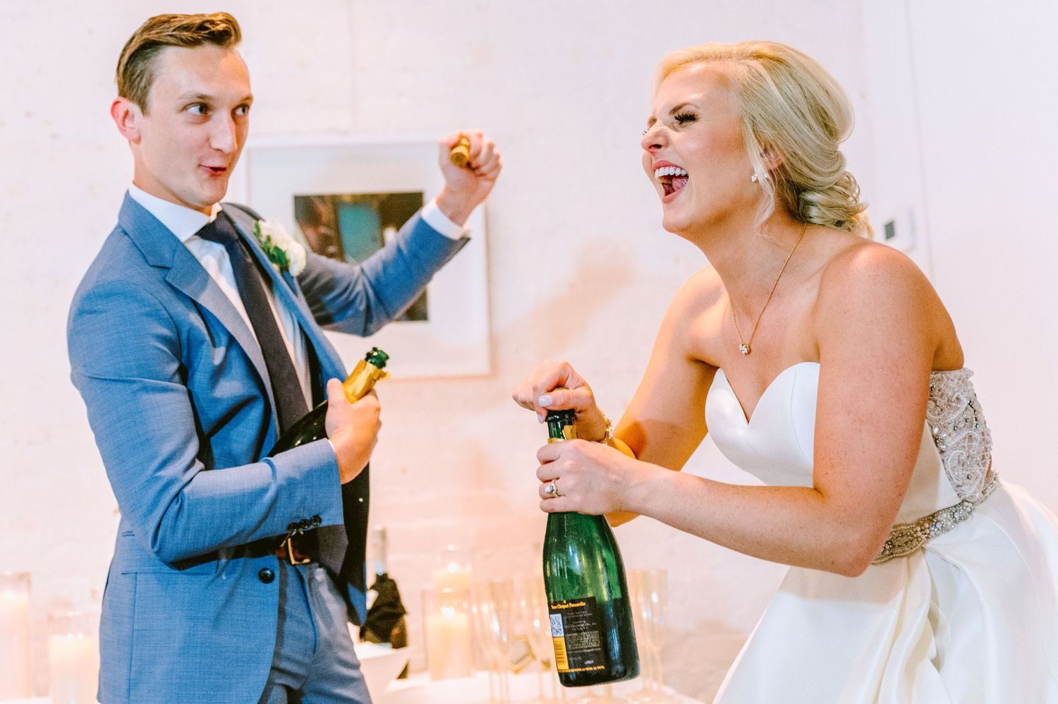A joyful couple in wedding attire celebrates by popping open champagne bottles together.